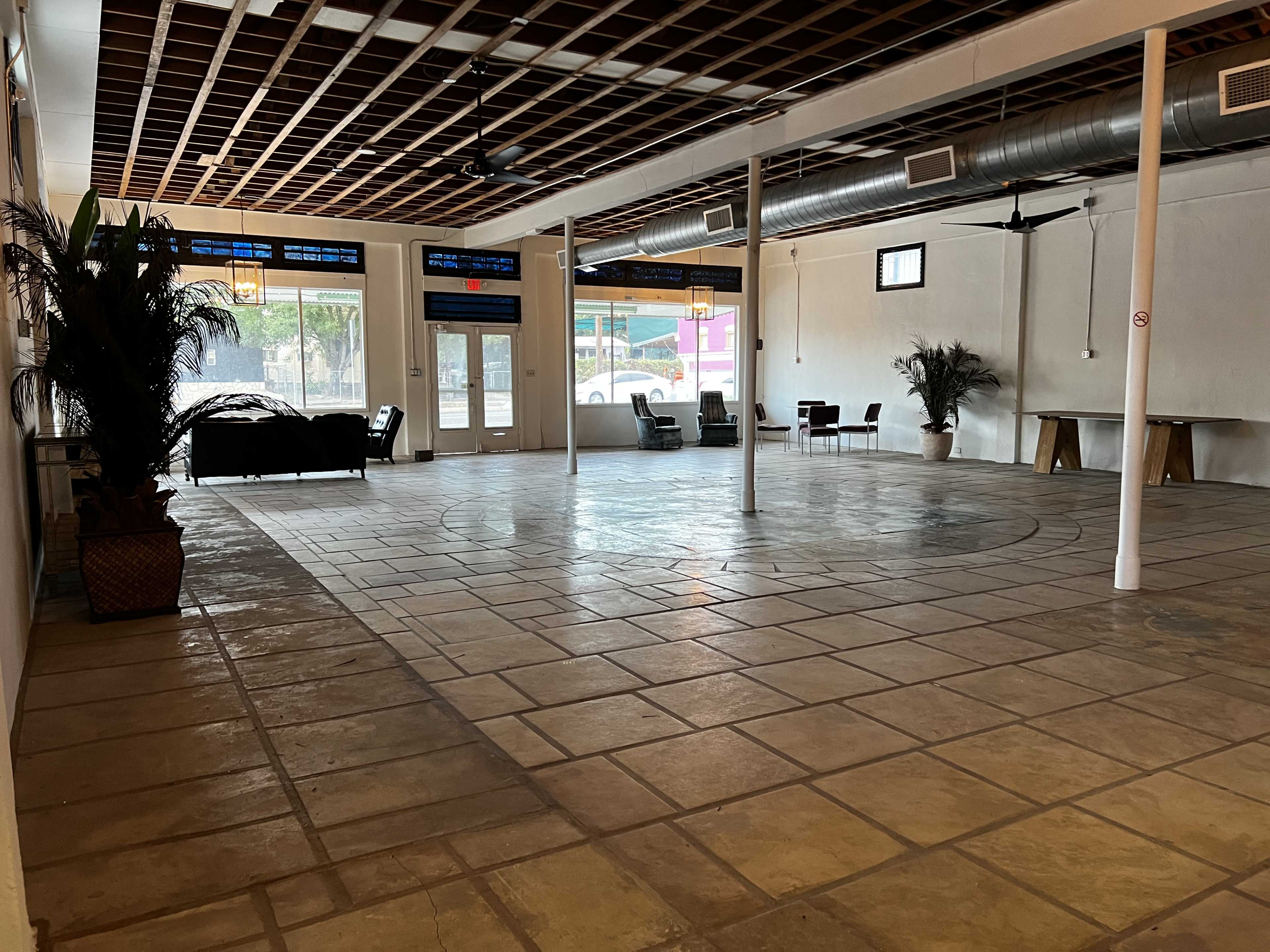 A spacious indoor room with tiled flooring, featuring a few chairs and a couch along the sides, and large windows letting in natural light.