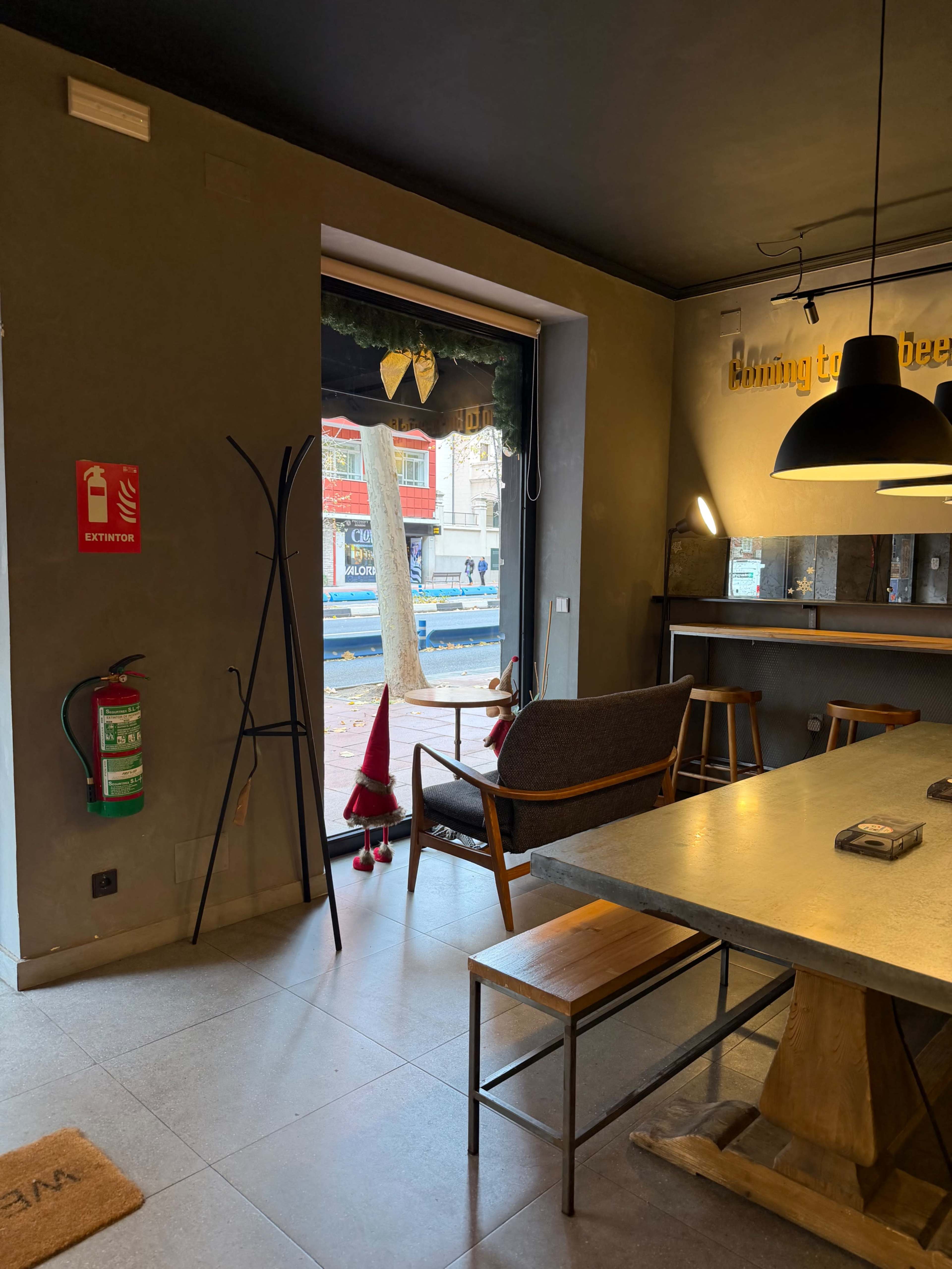 The image shows the interior of a café with a large window, a wooden table, and a chair, along with decorative elements and an exit sign.