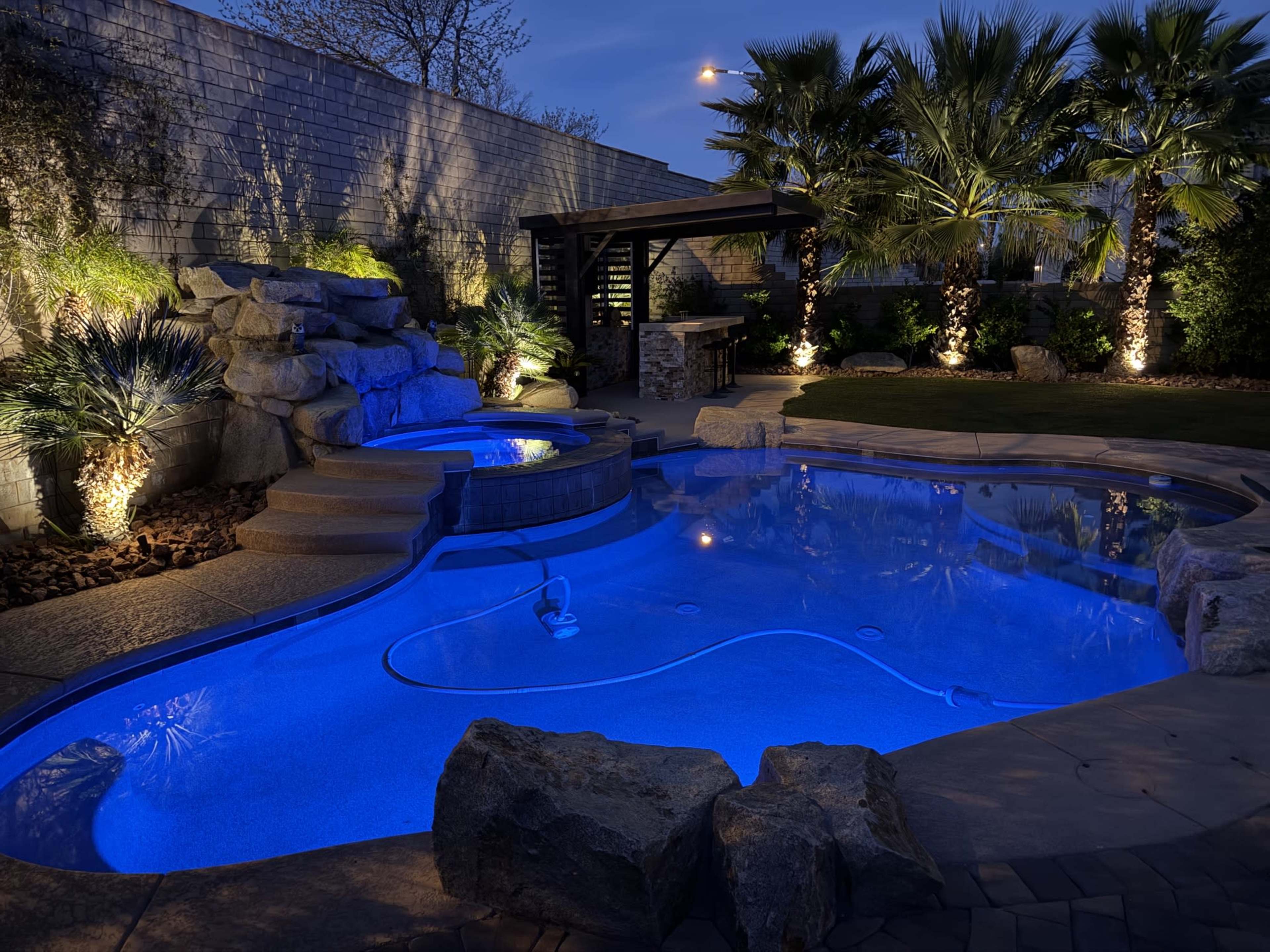A well-designed backyard features a pool with a spa, surrounded by landscaping and illuminated by soft lights at dusk.