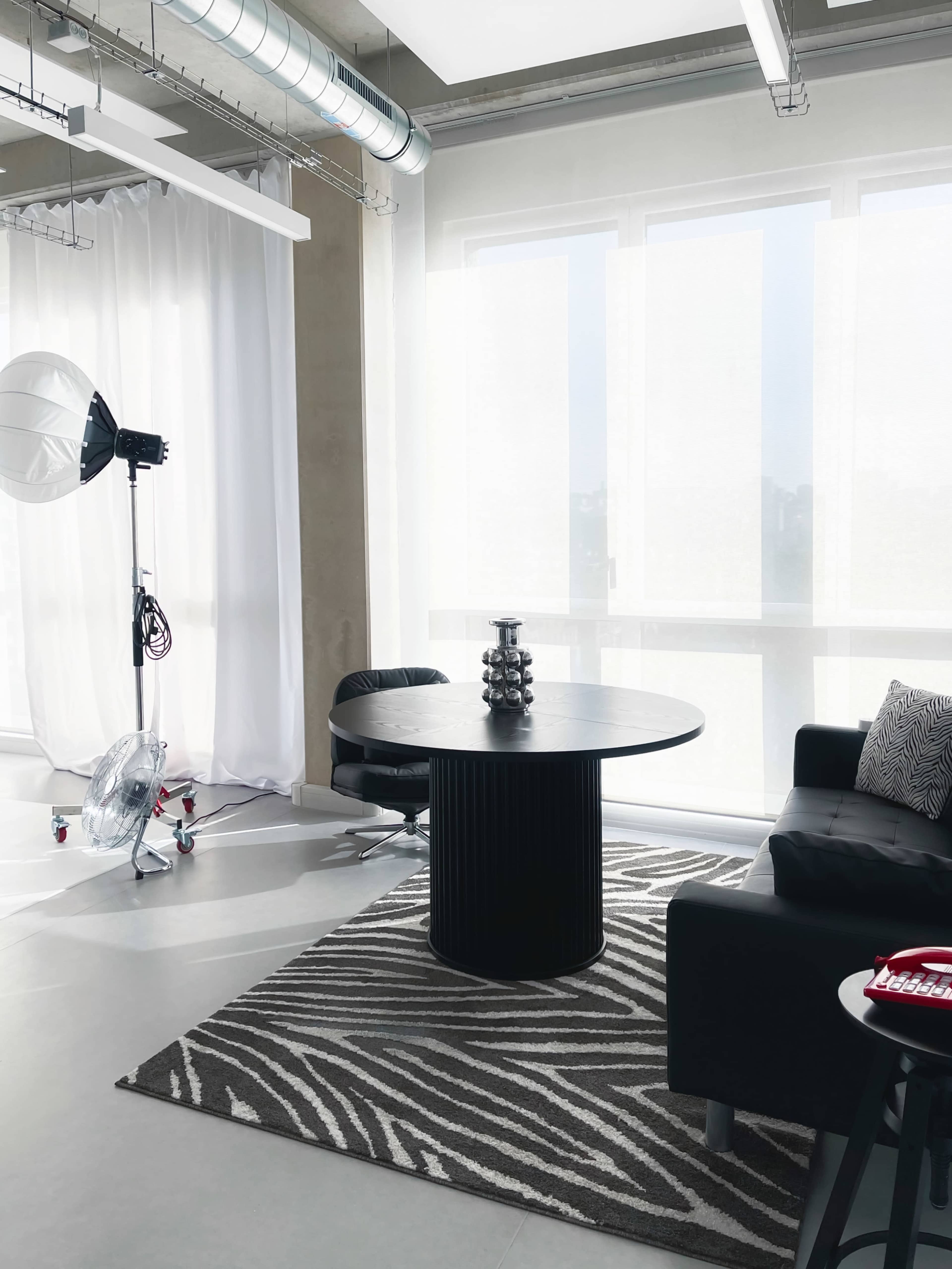 An interior room features a round black table, a black chair, and a couch, accompanied by a large photography light and sheer white curtains.