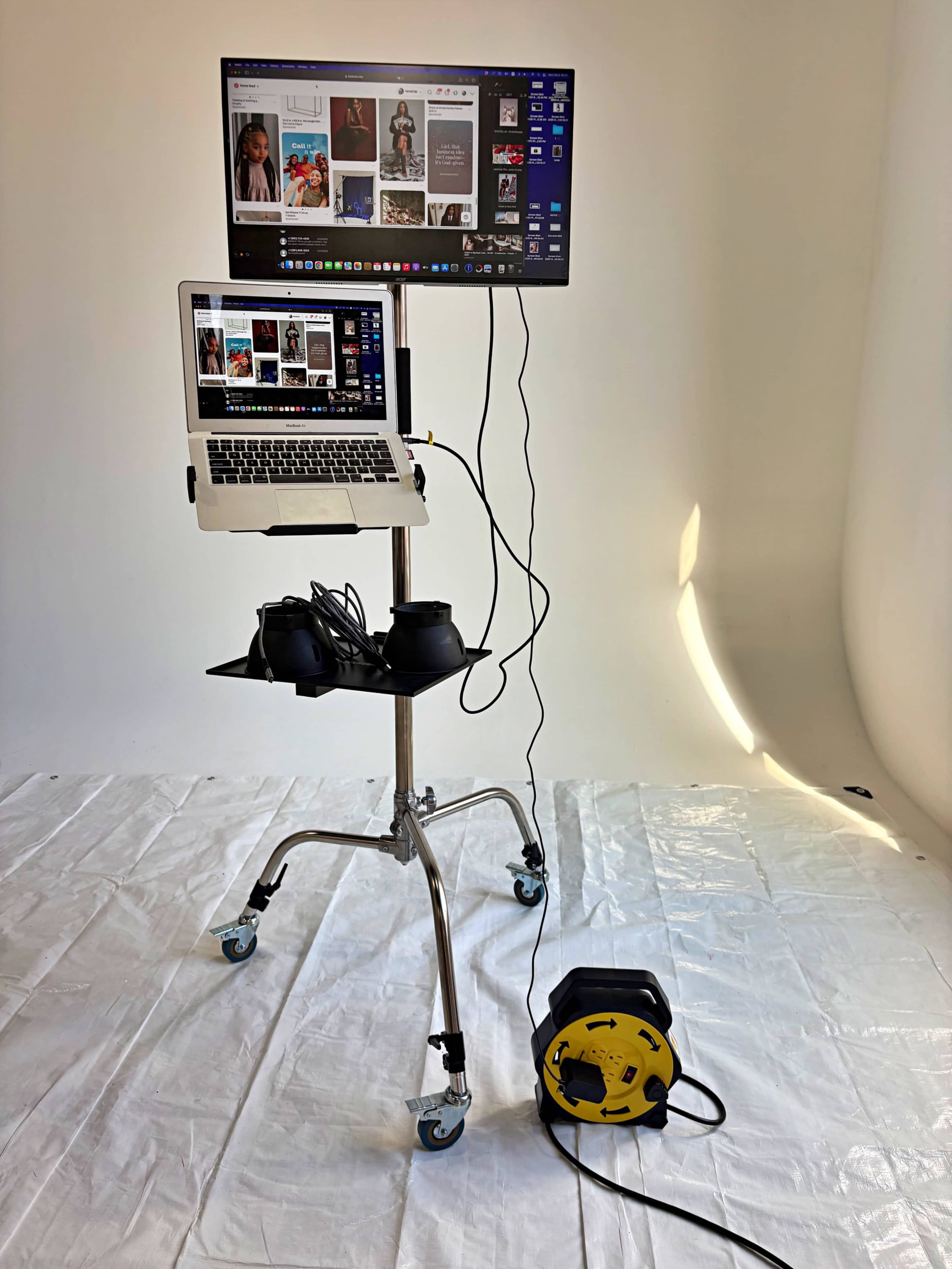 A laptop and external monitor are set up on a mobile stand, with VR headsets and a cable reel on the floor, against a plain background.