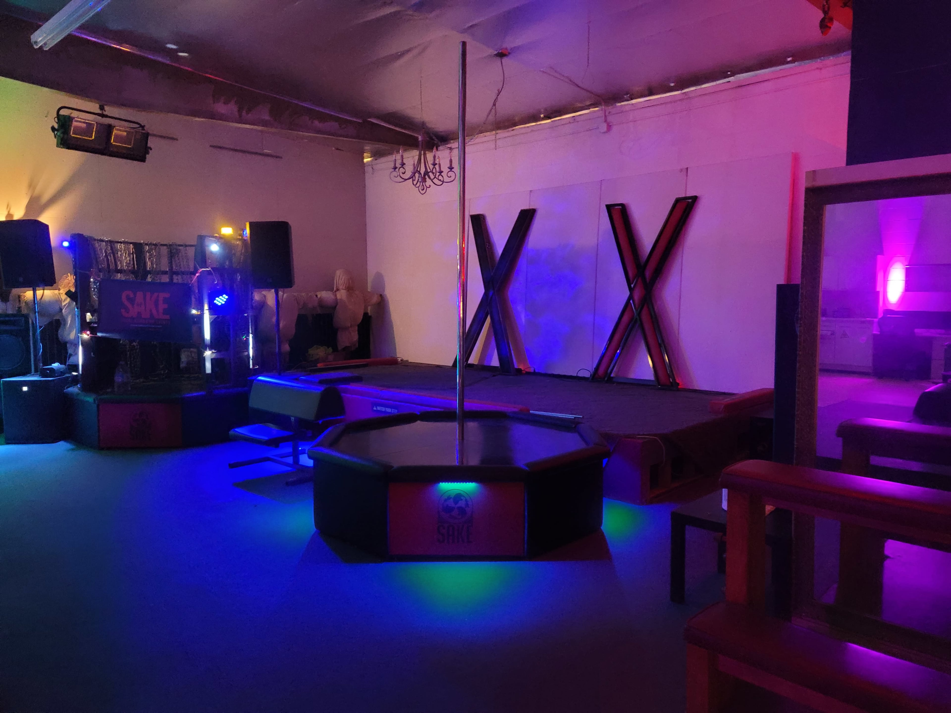A dimly lit entertainment space with a stage featuring a pole, surrounded by seating and illuminated by colorful lighting.