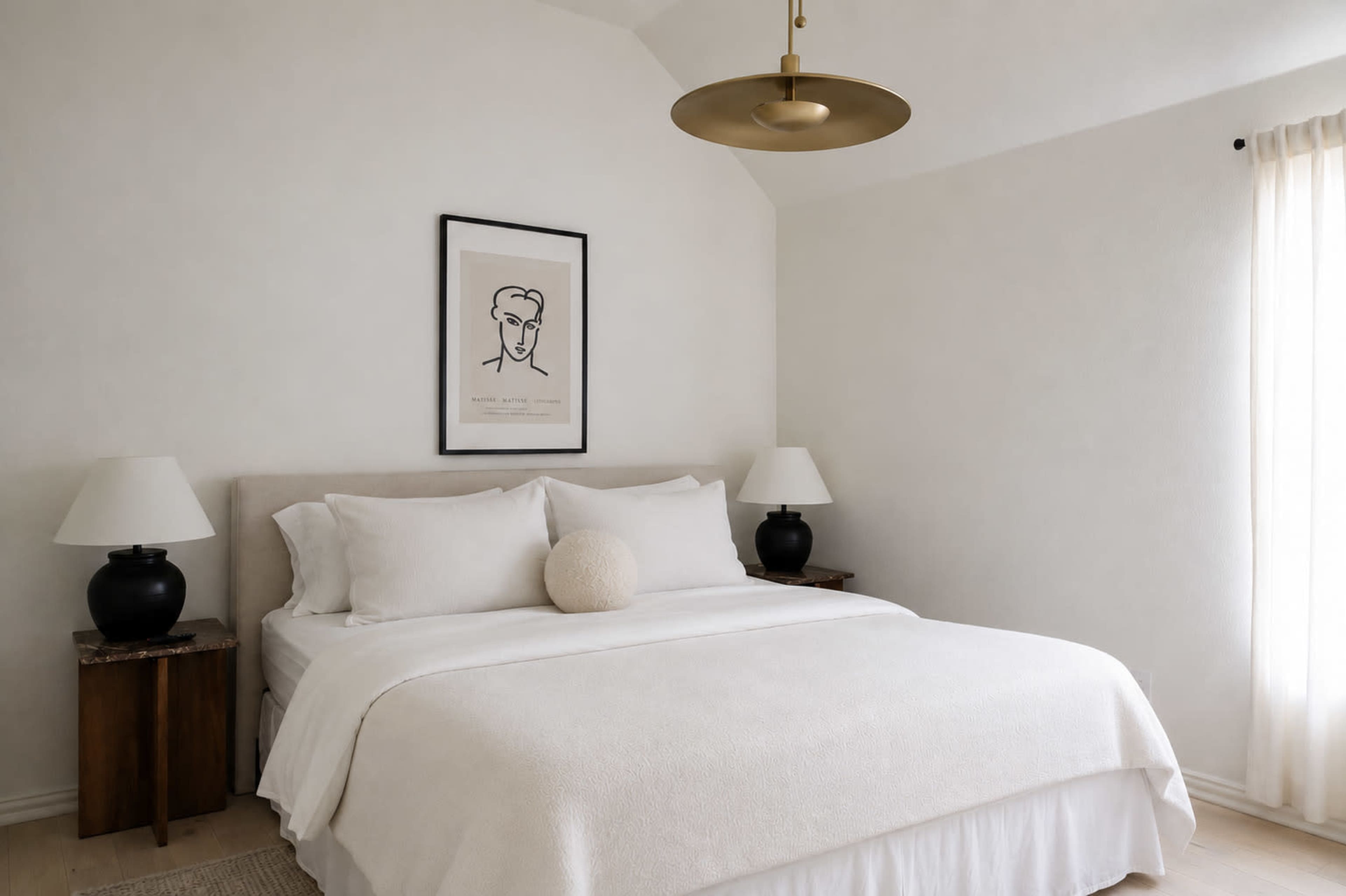 The image shows a minimalist bedroom featuring a large bed with white bedding, framed artwork on the wall, and two black lamps on wooden nightstands.