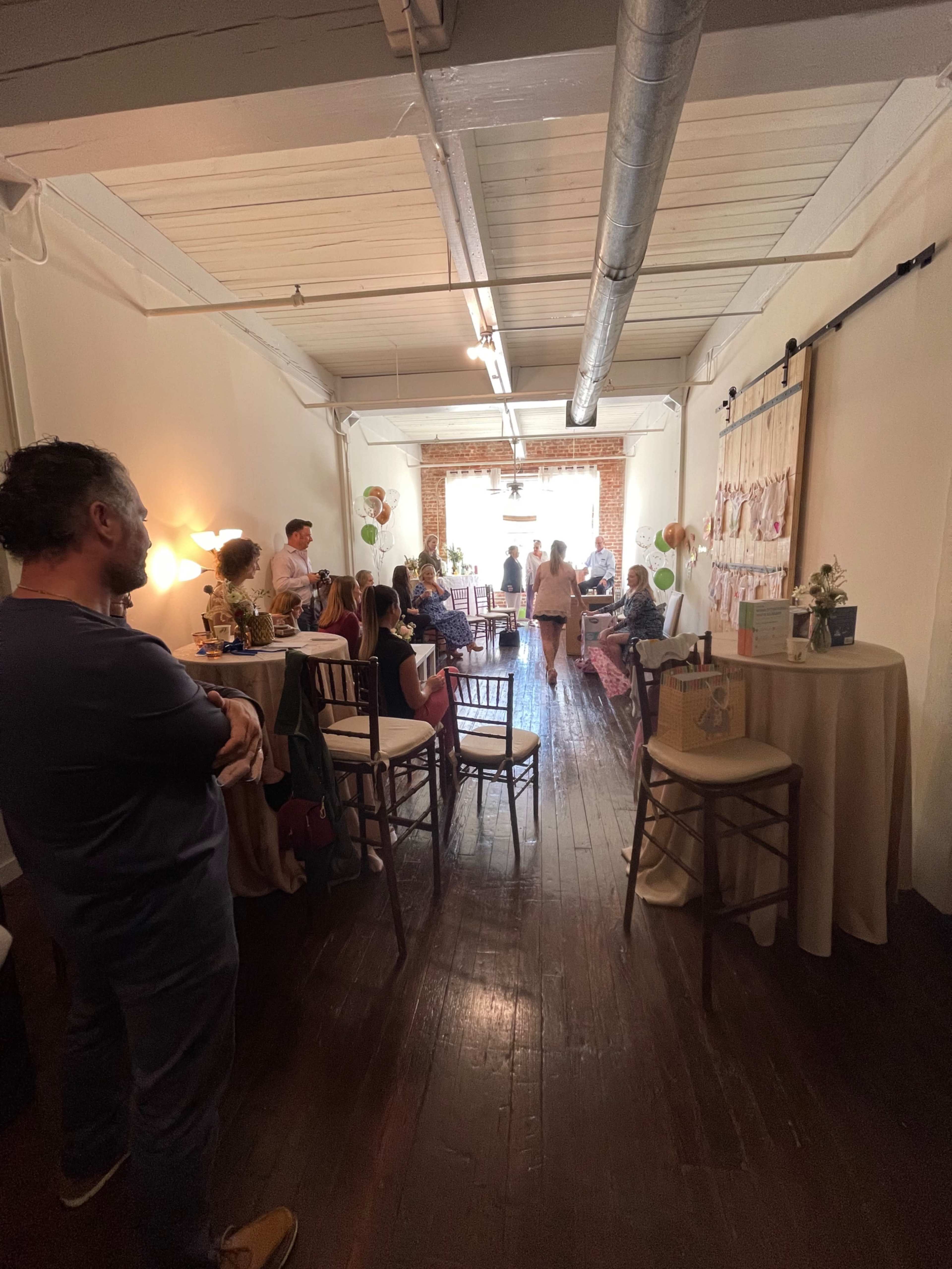 A group of people gathers in a spacious, warmly lit room with tables and decorations set up for an event.