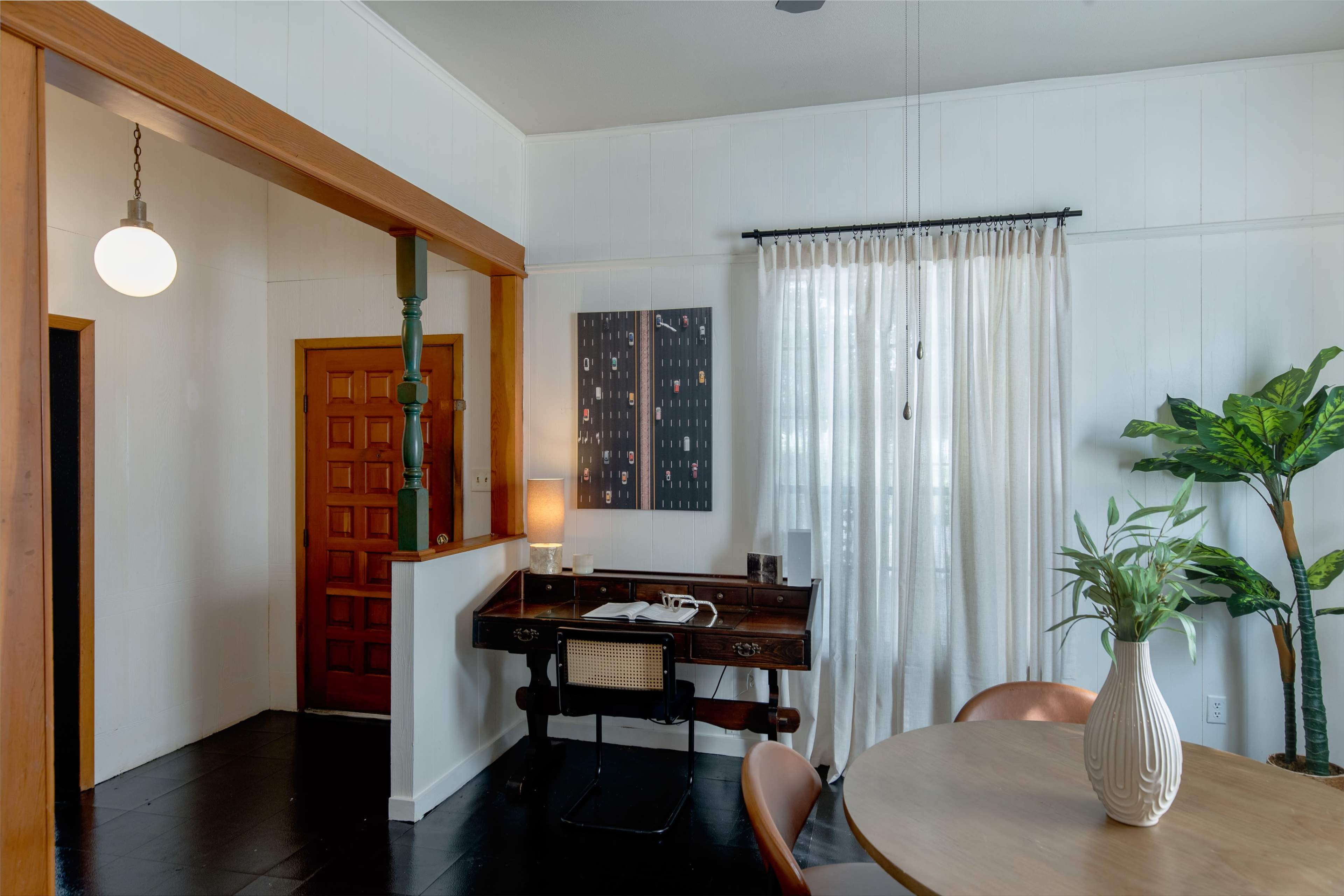The image shows an interior space featuring a small desk and chair with a lamp, a round dining table, a potted plant, and a large window with curtains, alongside a wooden door and entryway.