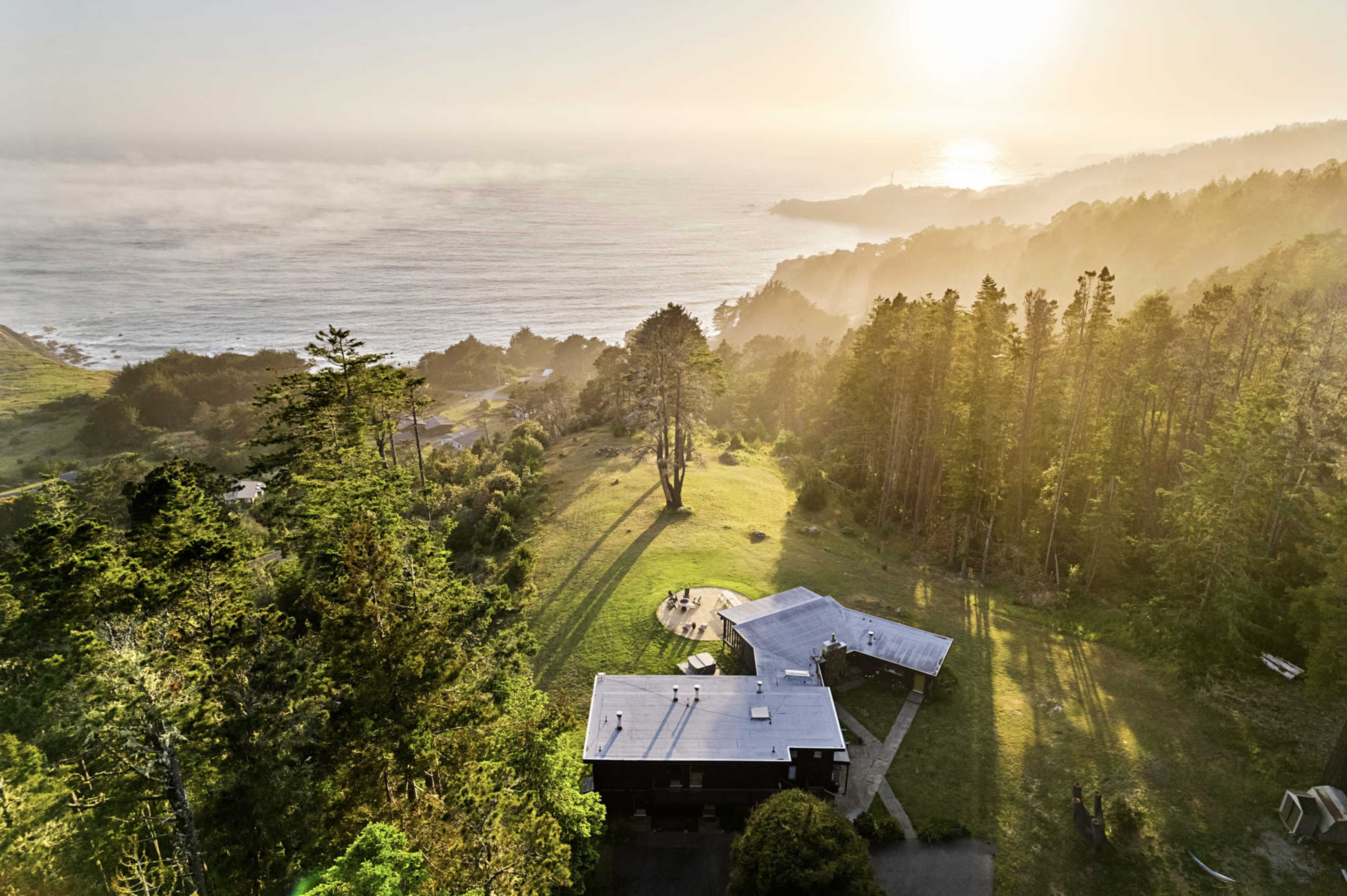 The image shows a wooden house situated on a grassy area overlooking the ocean, surrounded by trees and hills at sunrise.