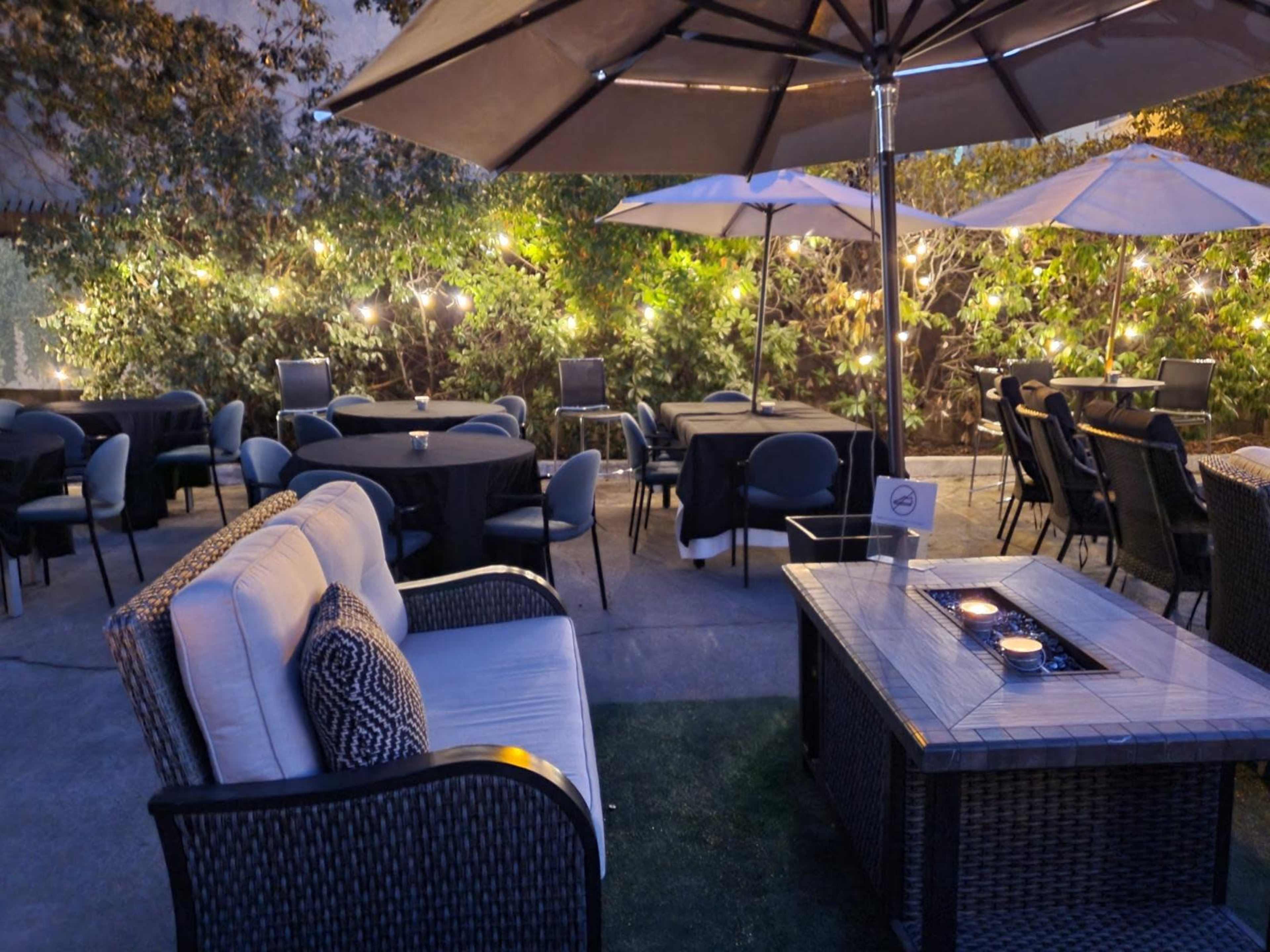 A cozy outdoor seating area with tables, chairs, and umbrellas surrounded by greenery and softly glowing lights.