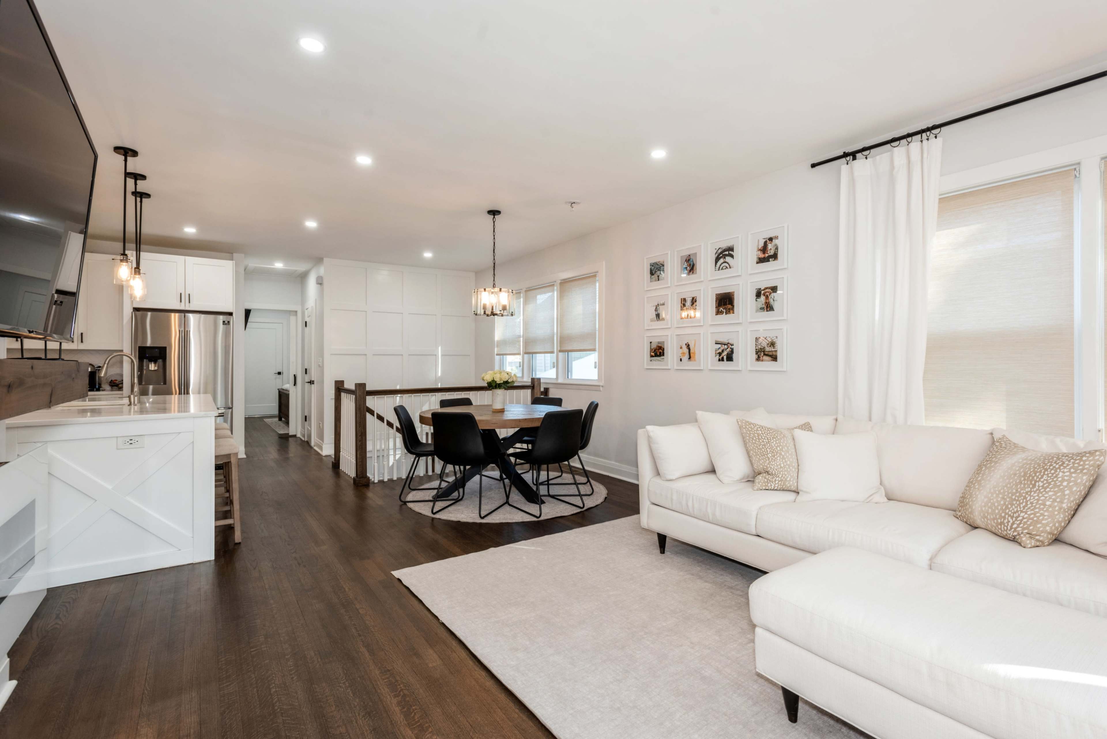 A modern open-concept living space featuring a white sectional sofa, a dining area with a round table and black chairs, and a kitchen with white cabinetry.