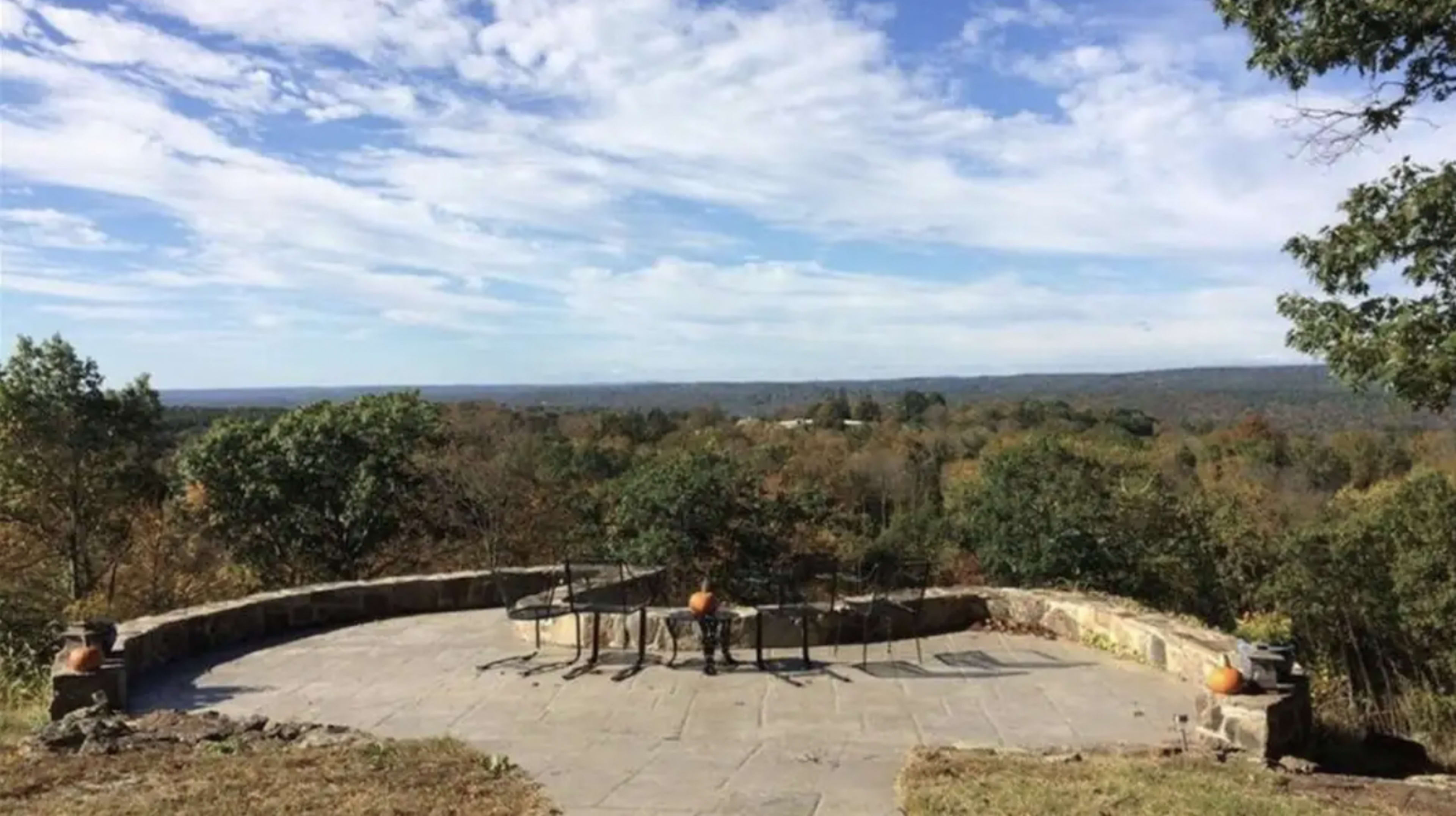 A stone patio with a circular table and chairs overlooks a valley filled with trees under a partly cloudy sky.