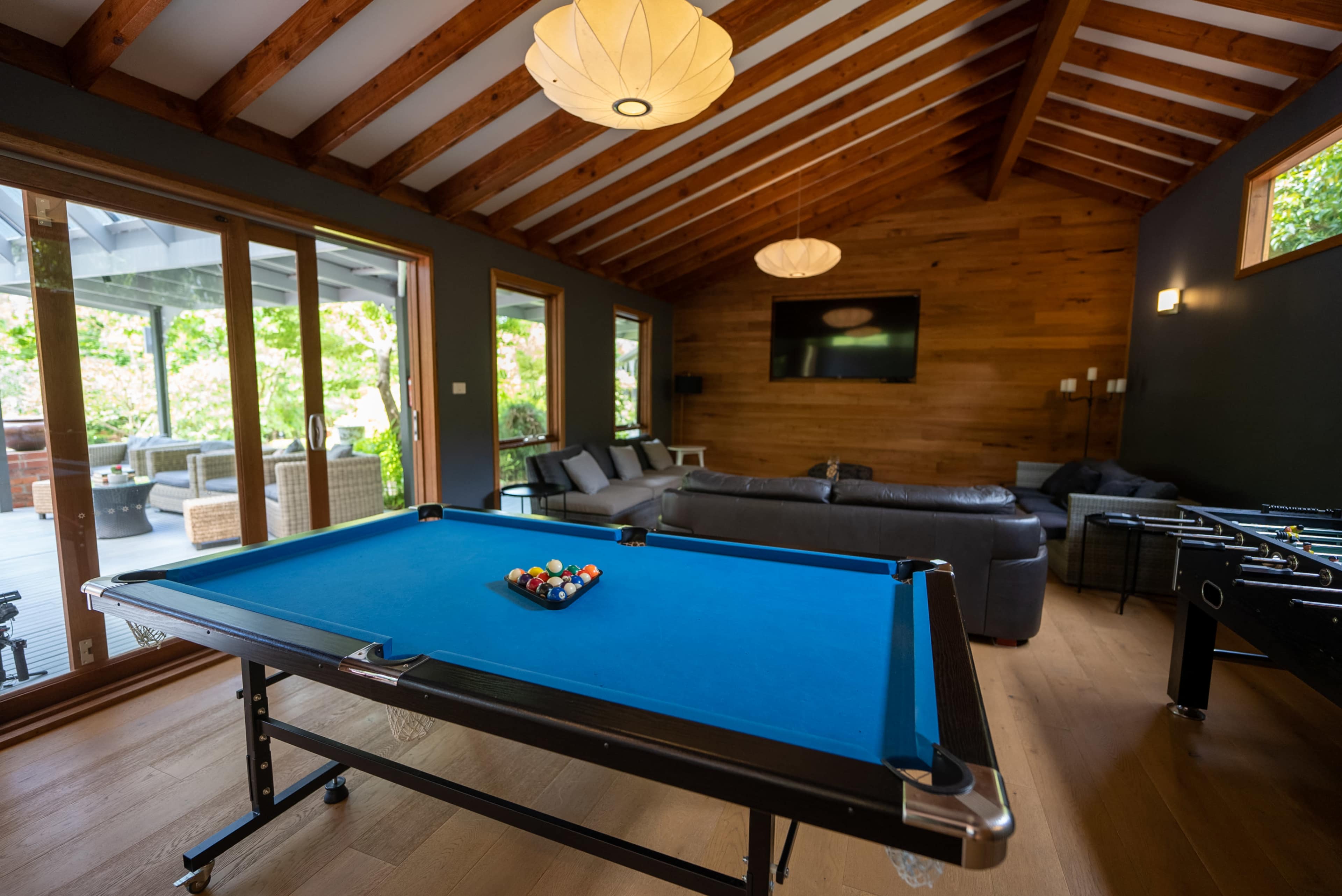 A blue pool table is positioned in a spacious room with wooden beams, surrounded by large windows and a seating area.