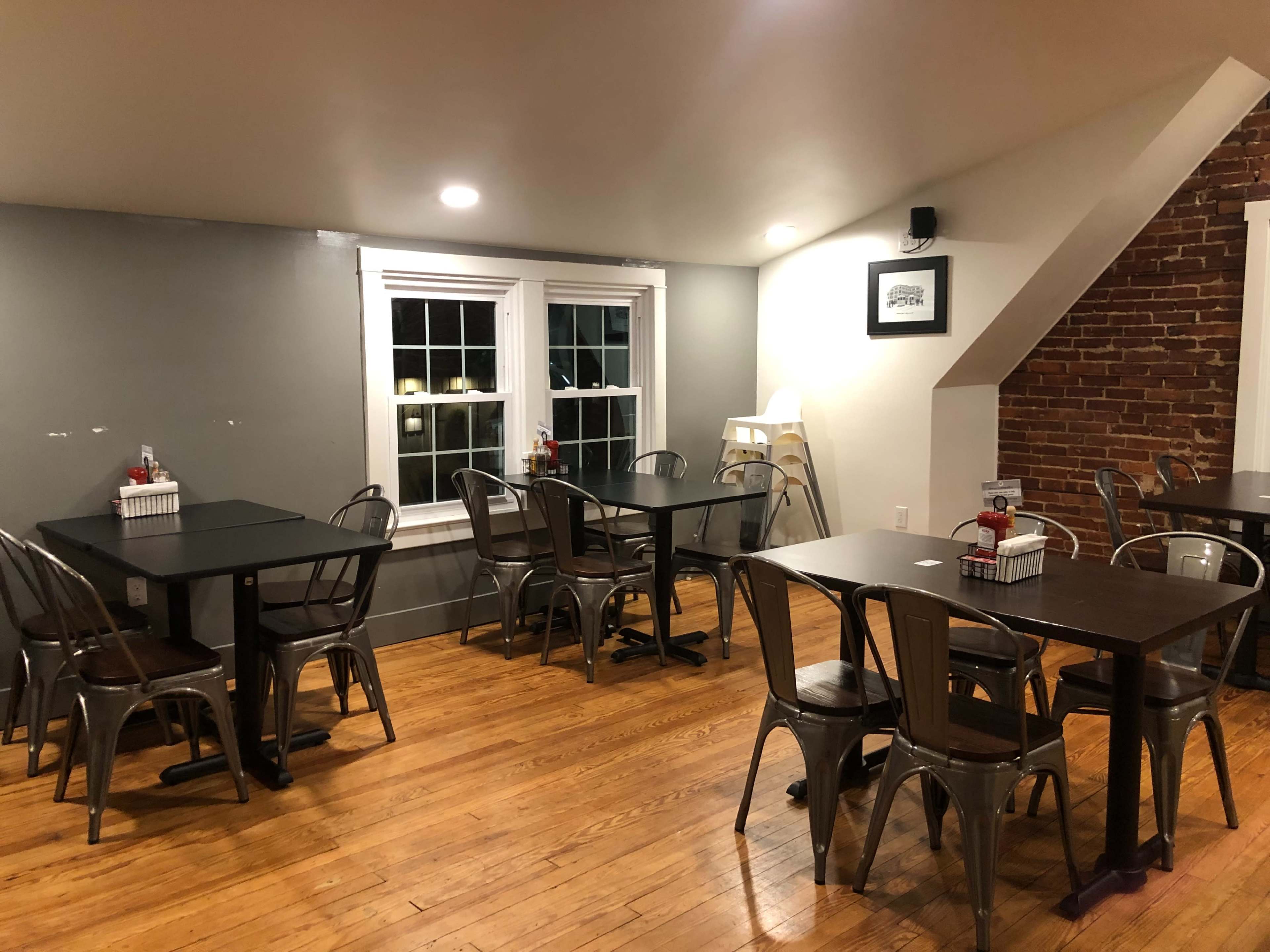 The image shows an open dining area with tables and chairs arranged, featuring a brick wall and large windows with a view of the outside.