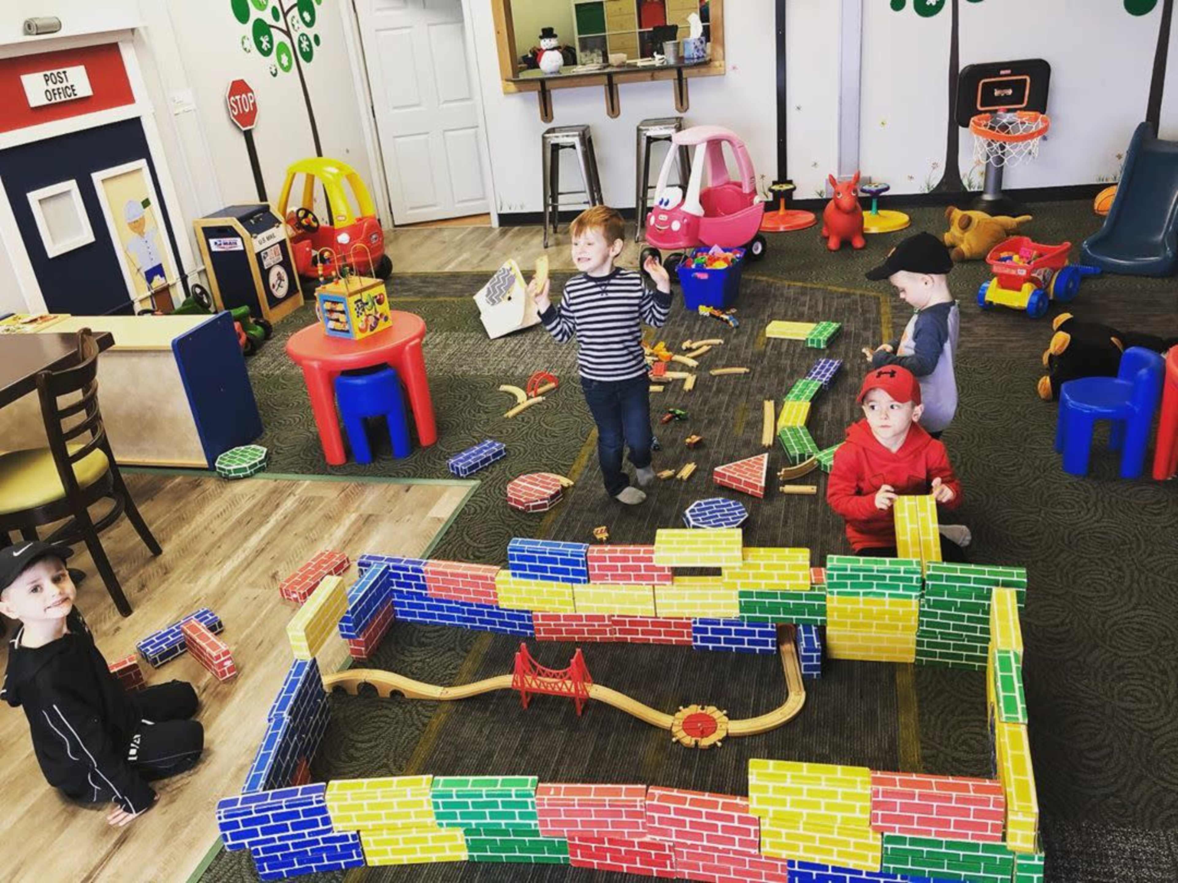 Four children are building a colorful block structure on a playmat in a room filled with various toys and play equipment.