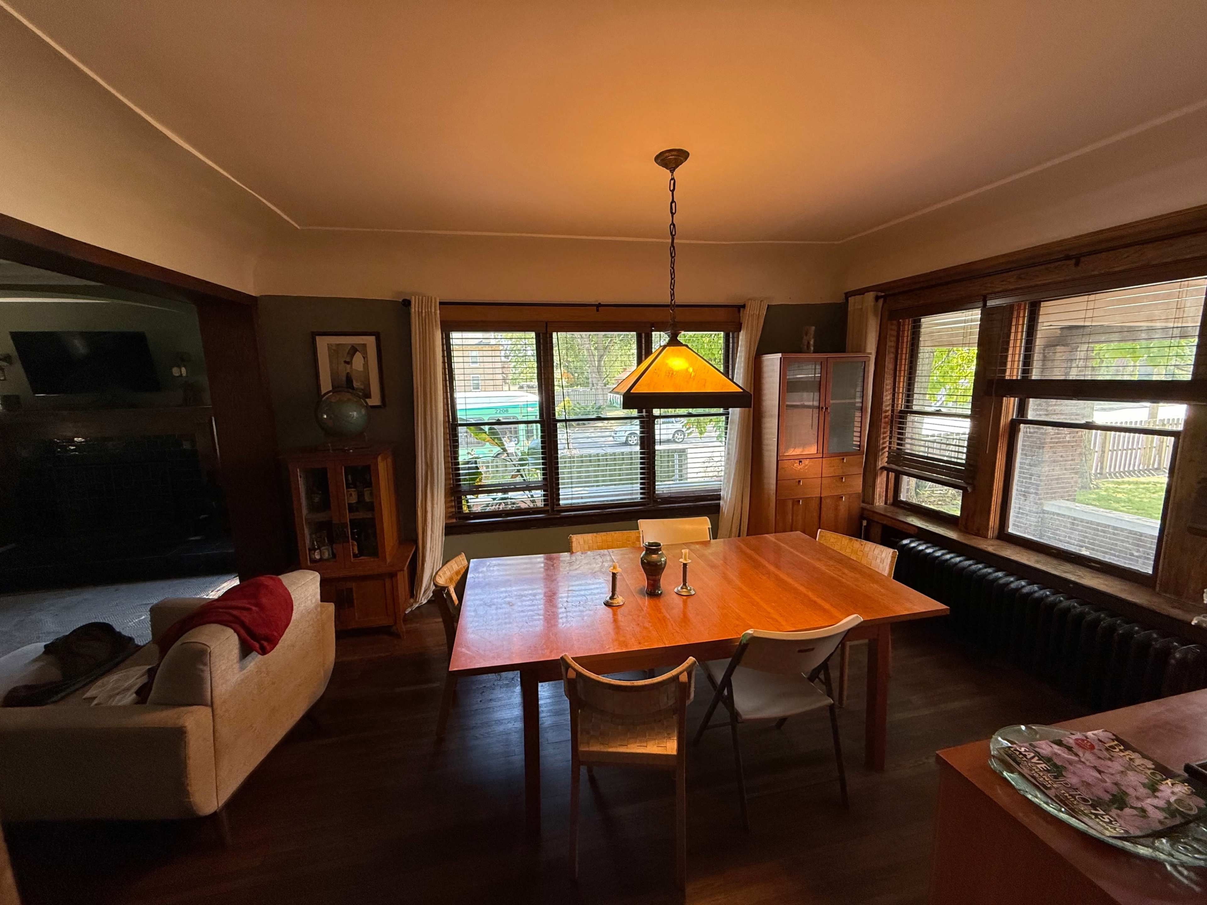 A dining room features a wooden table with two candles, surrounded by chairs, and illuminated by a hanging light fixture, with large windows offering views of the outdoors.