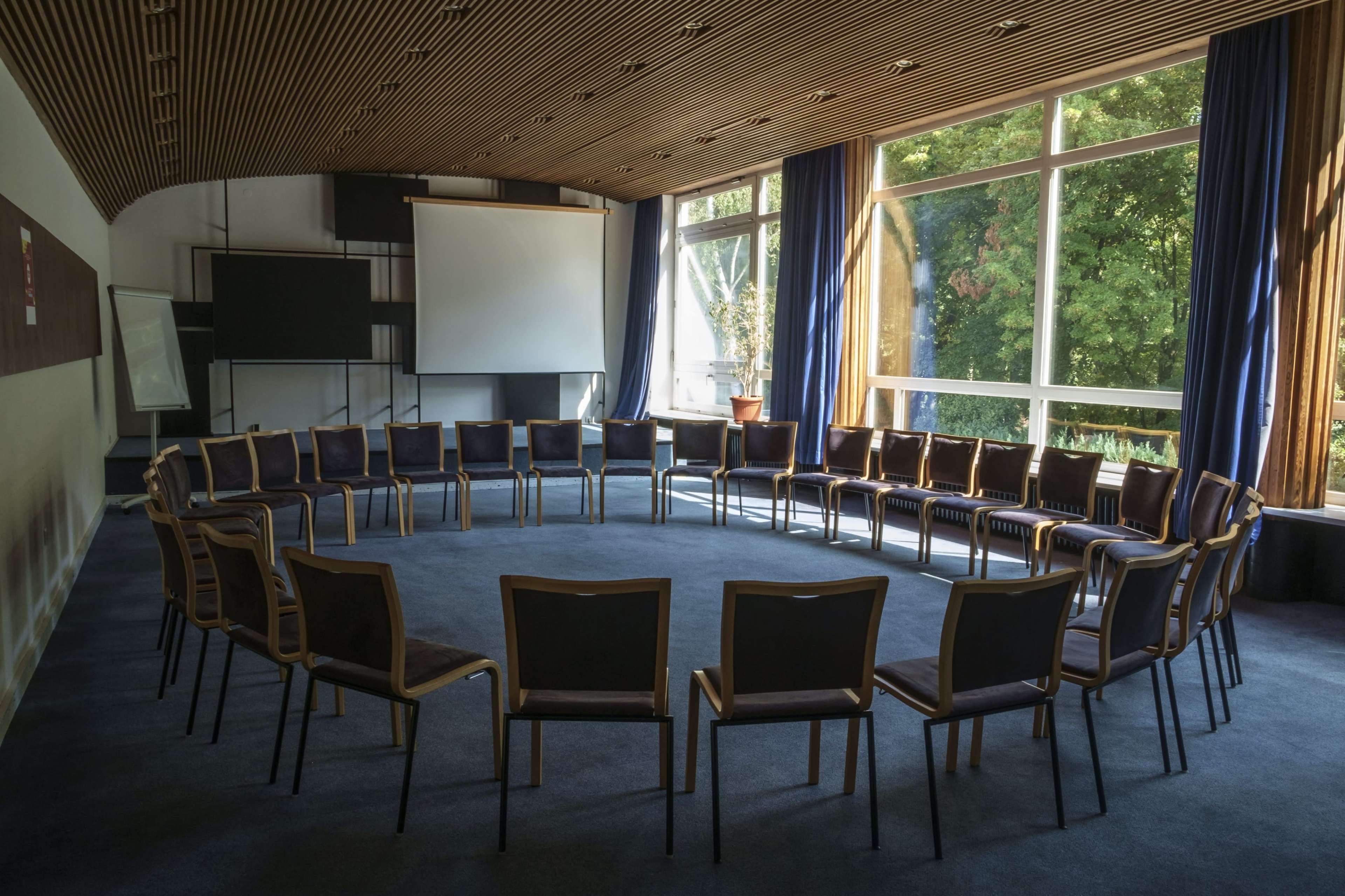 A circular arrangement of wooden chairs is set up in a well-lit conference room with large windows and a projector screen.
