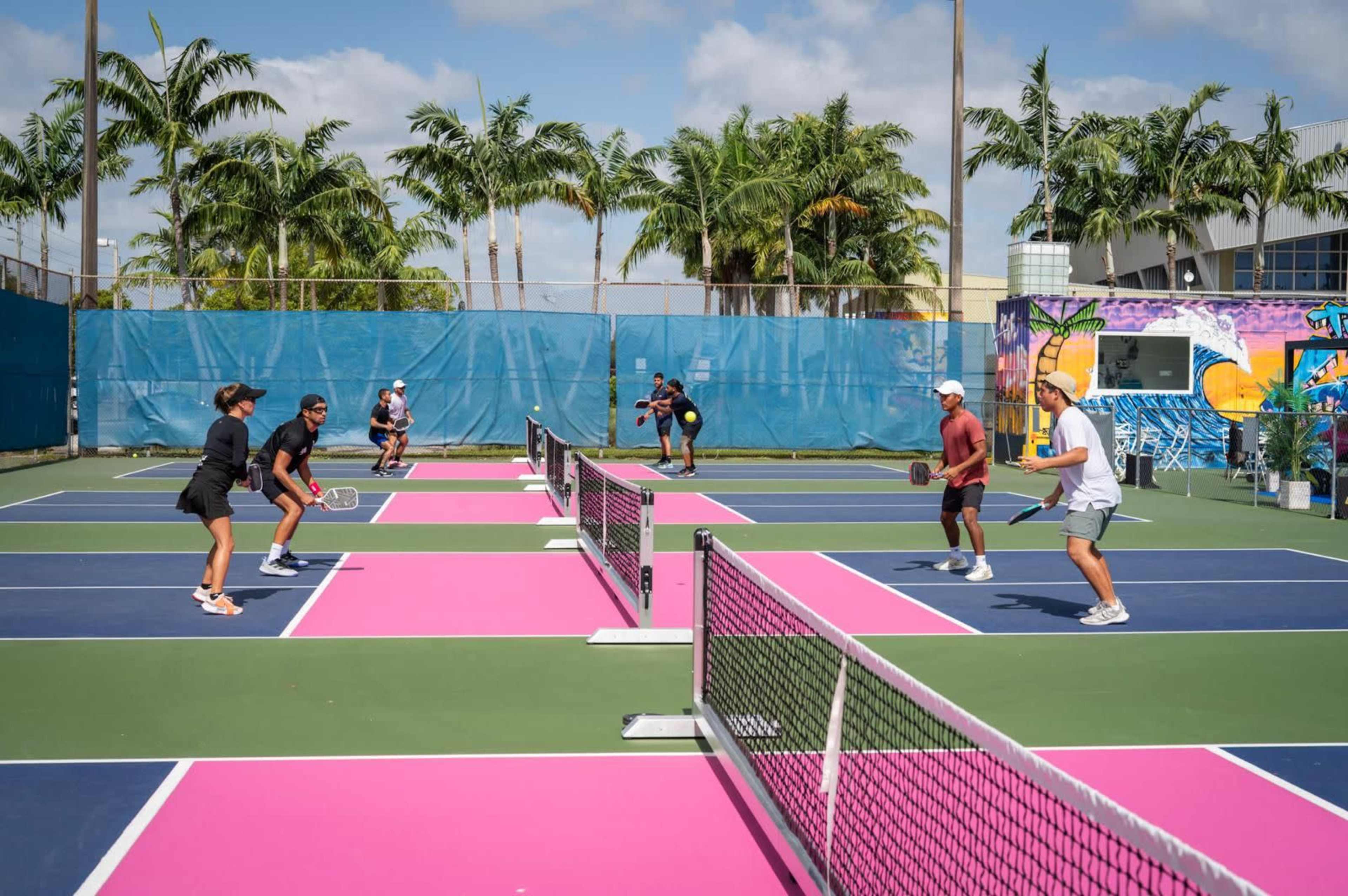 Two pairs of players engage in a pickleball game on a vibrant court with pink and blue sections, surrounded by palm trees and an urban mural.
