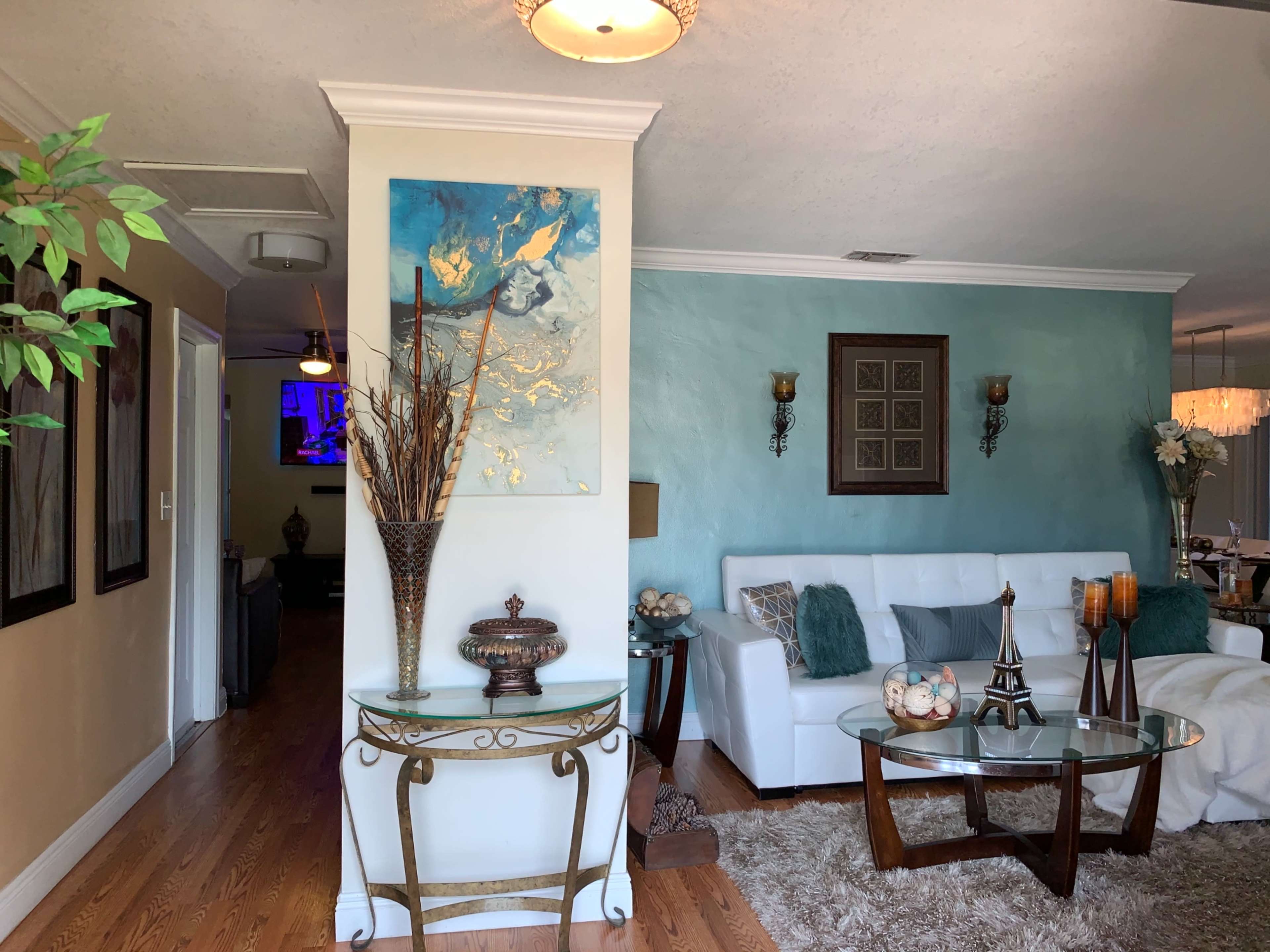 The image shows a modern living room adorned with a white sofa, decorative wall art, and a glass coffee table surrounded by a stylish rug.