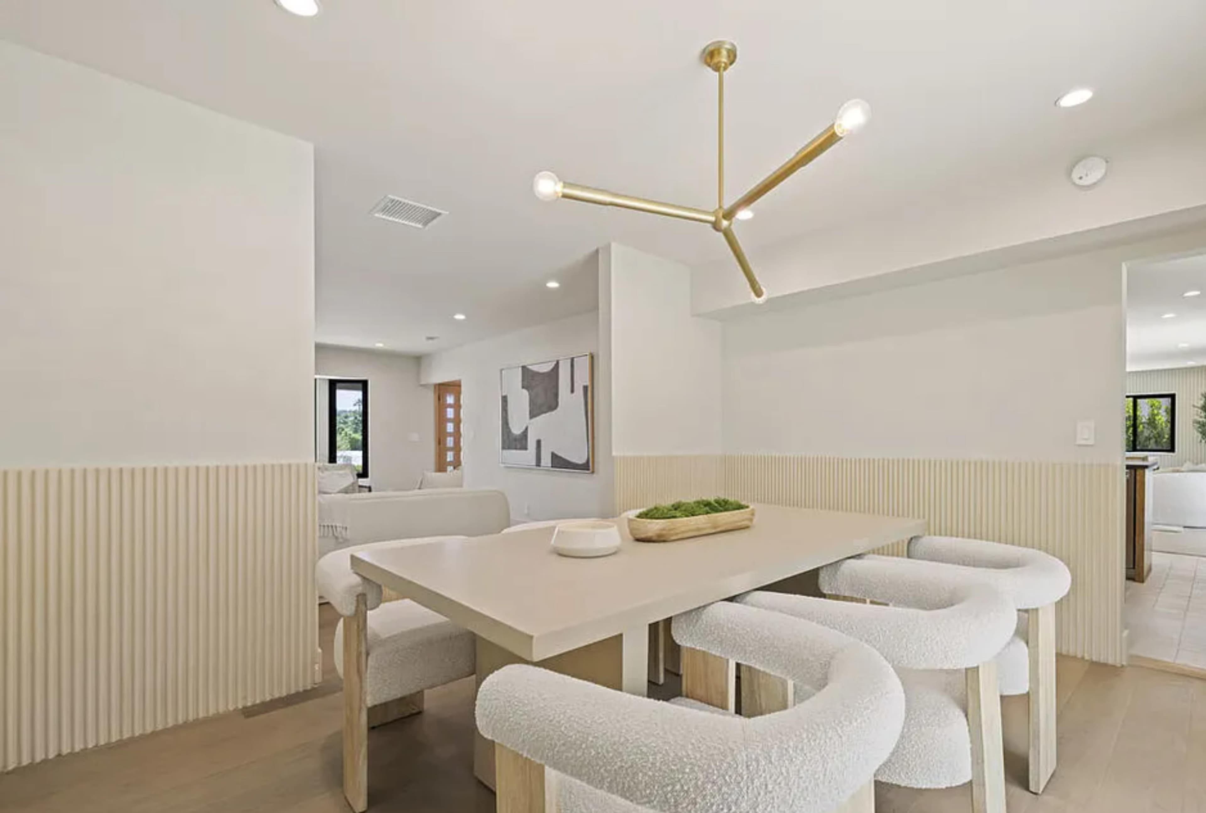 The image shows a modern dining area featuring a light-colored table surrounded by four upholstered chairs and a geometric pendant light overhead.
