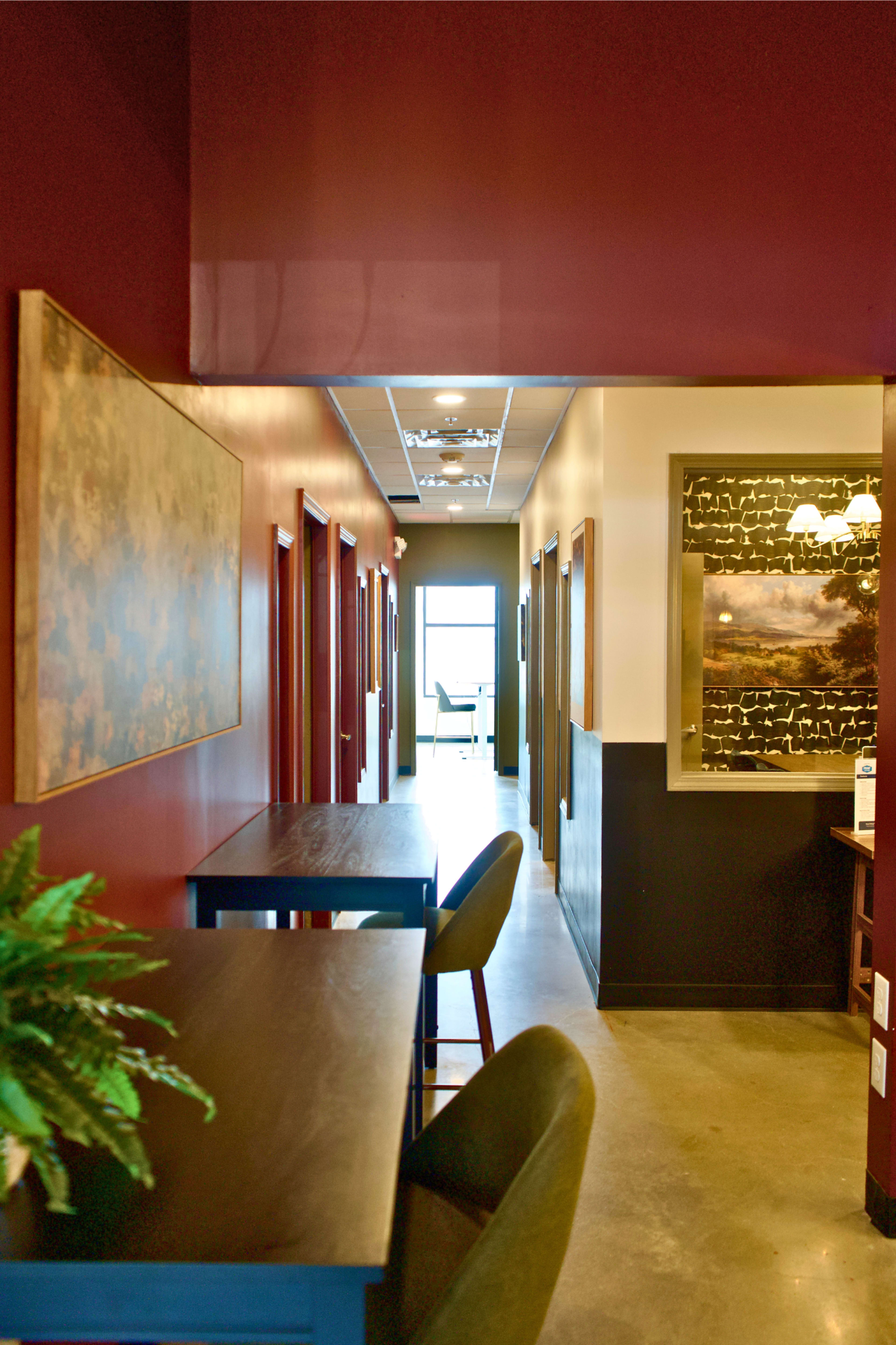 The image shows a hallway in a building with lined doors, artwork on the walls, and a table with chairs on one side.