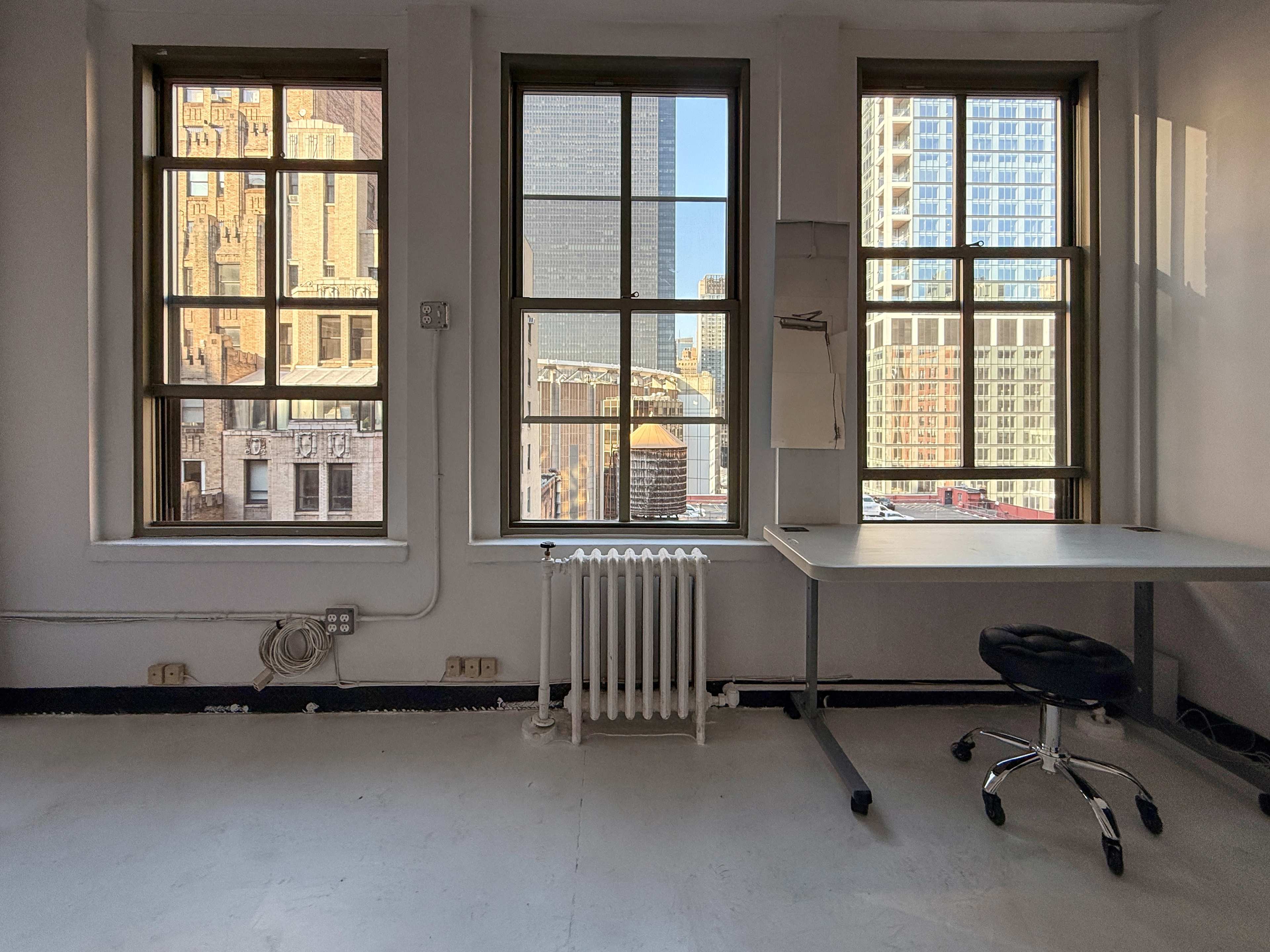 The image shows a vacant office space with three large windows, a metal radiator, and a simple desk with a rolling chair.