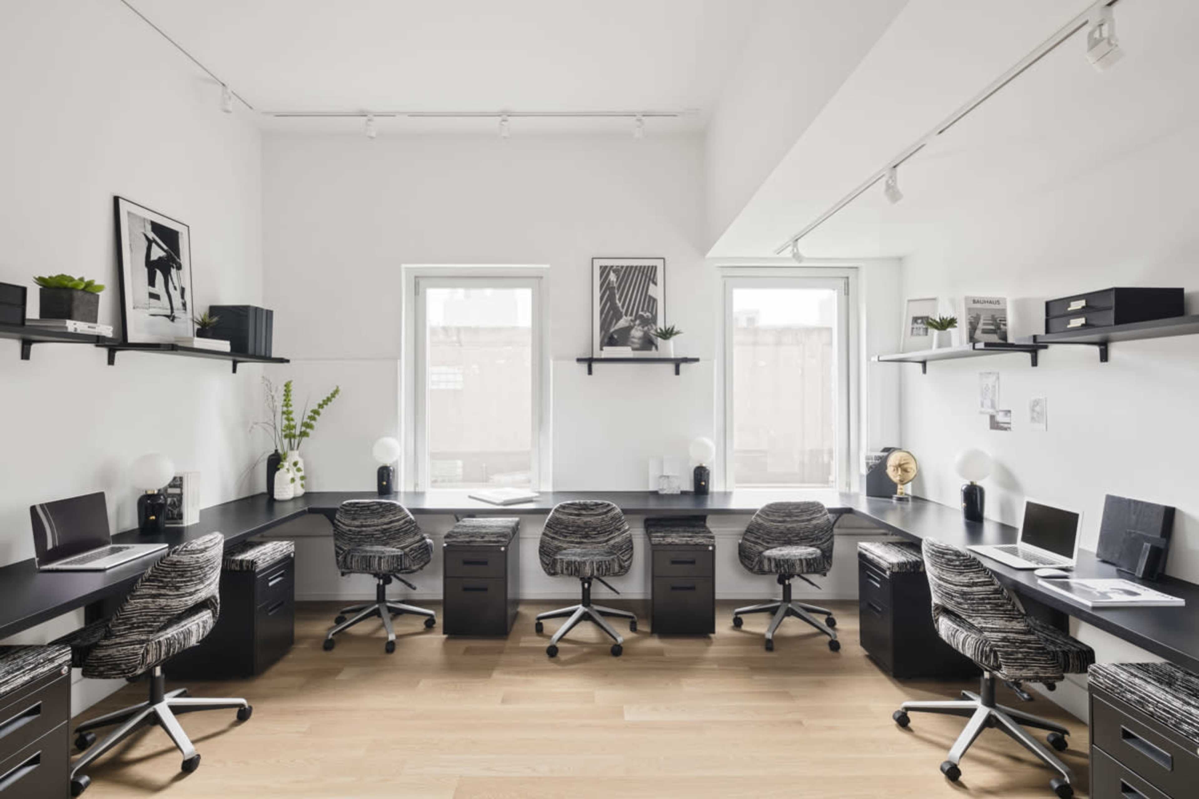 A modern office space features four ergonomic chairs around a U-shaped black desk with shelves holding decorative plants and framed artwork.
