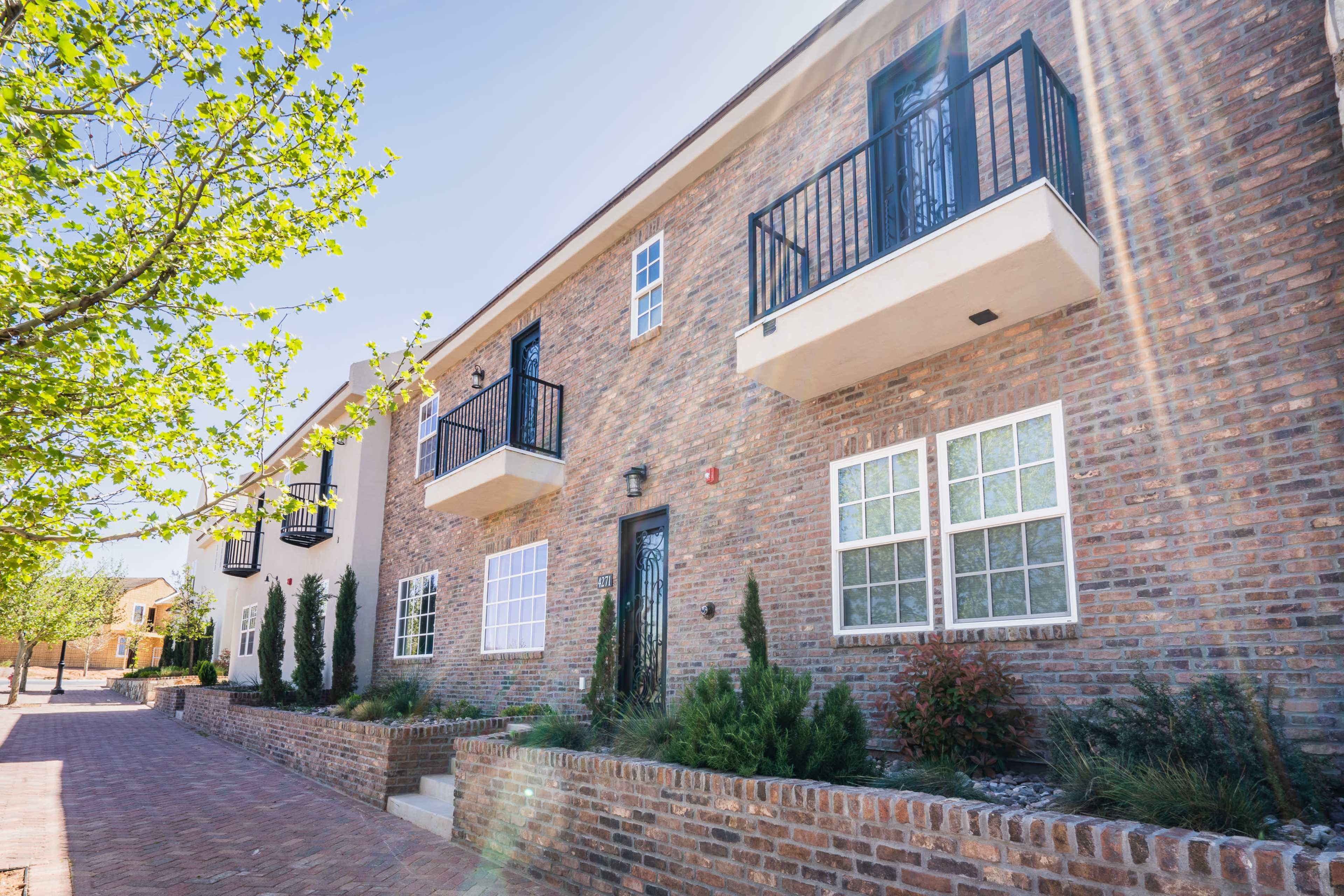 A row of brick townhouses with balconies, framed by trees and a paved walkway.