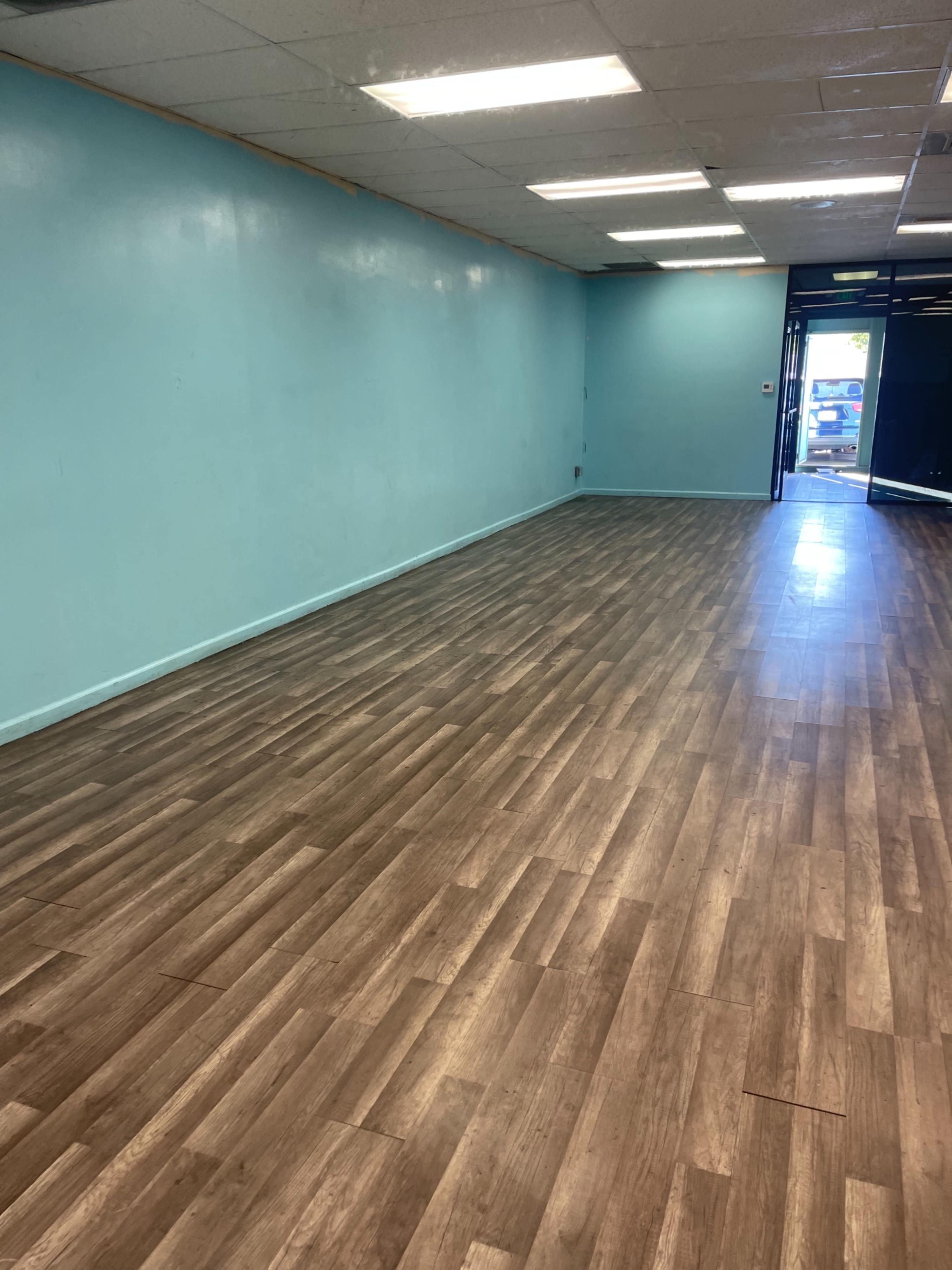 A spacious interior room with light blue walls and a wooden floor, featuring a glass entrance door at one end.
