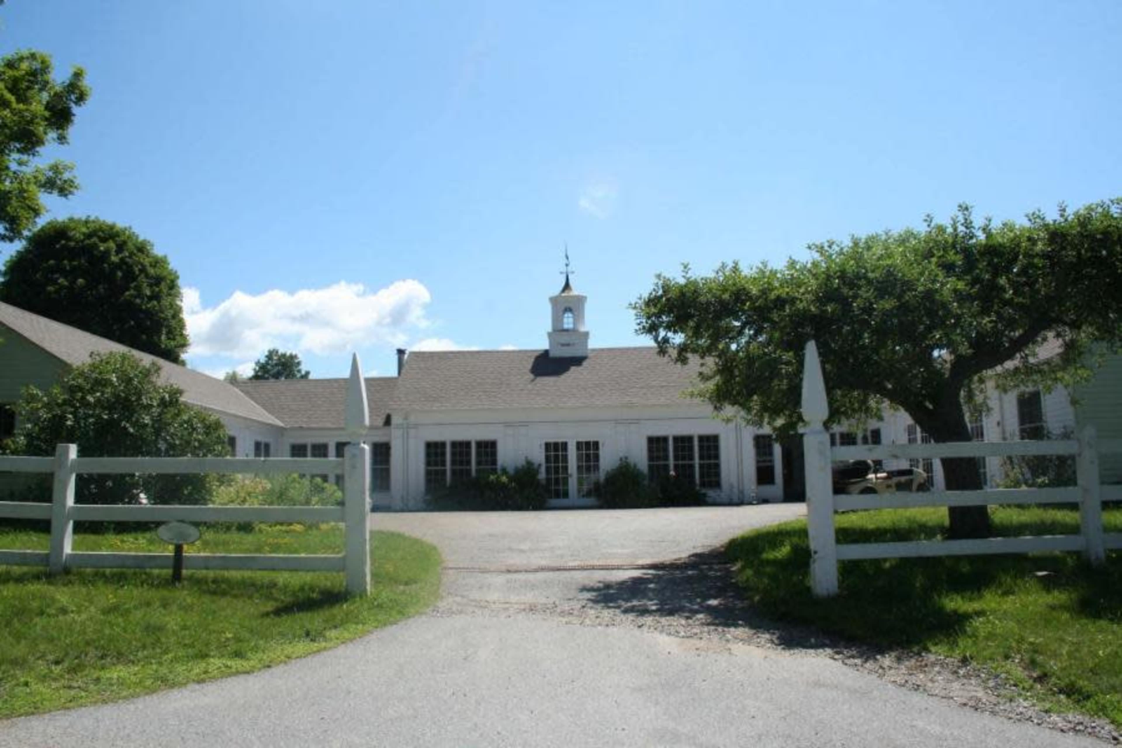 A white building with a steeple is surrounded by green trees and a gravel driveway, leading to a fenced entrance.