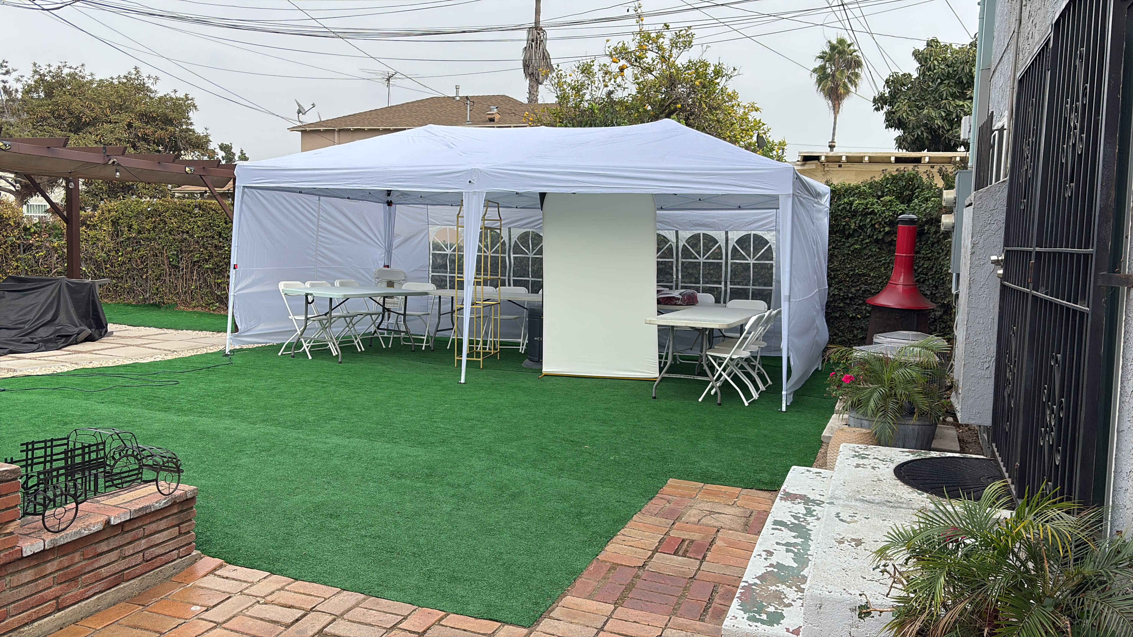 A white canopy tent is set up in a backyard with green artificial grass, featuring several white tables and chairs inside.
