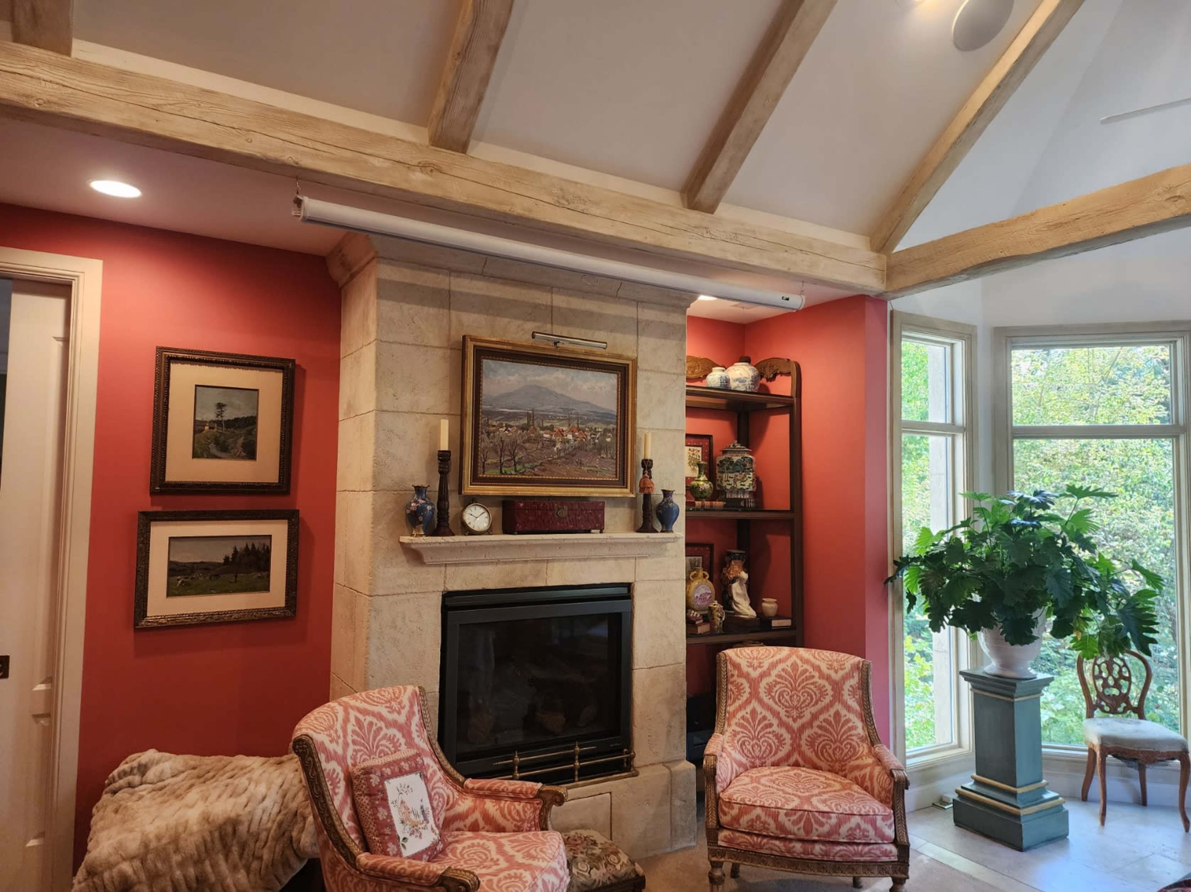 The image shows a cozy living room with a stone fireplace, decorative shelves, a large painting, and two patterned armchairs facing the fireplace.