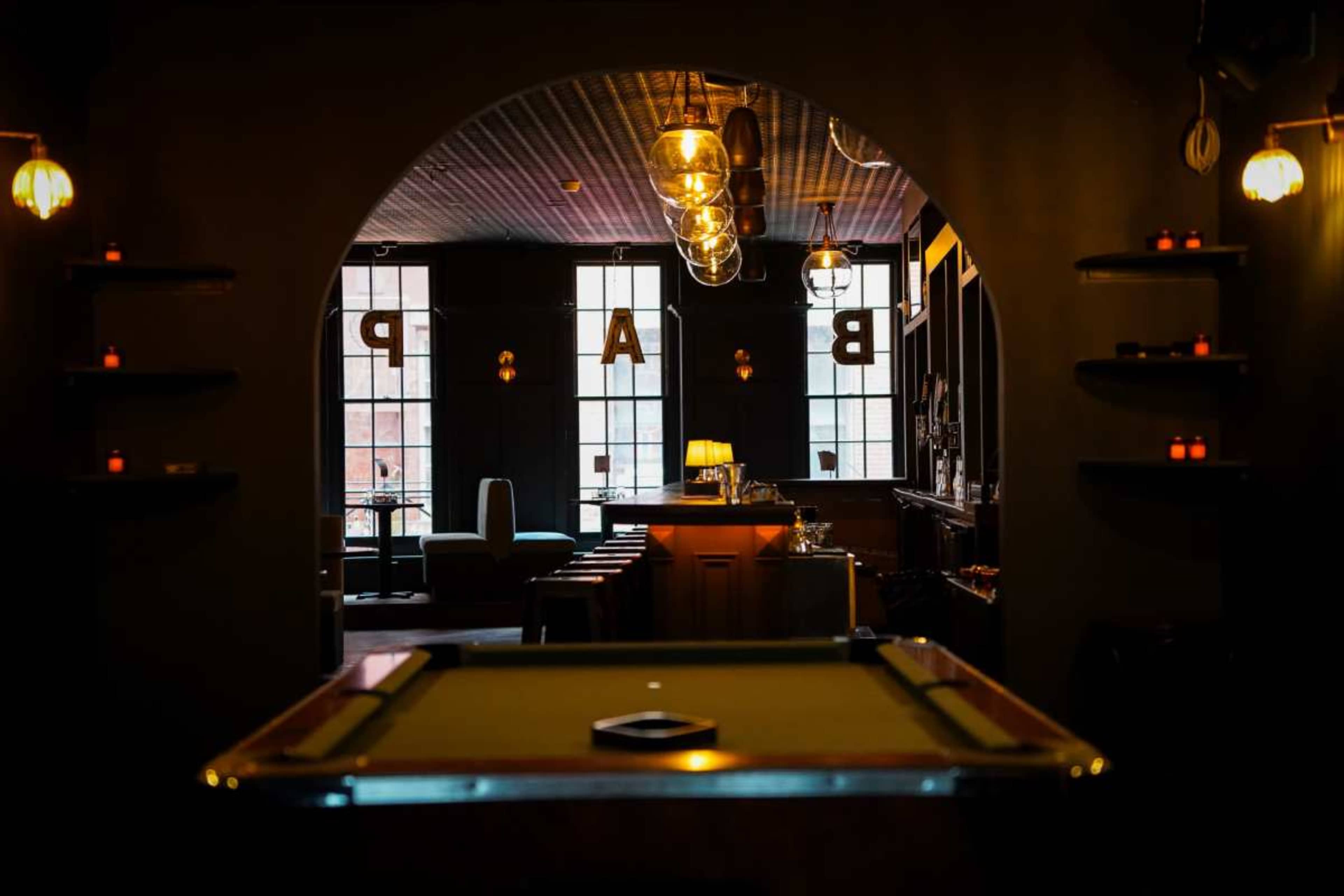 The image shows a dimly lit bar area with a pool table in the foreground and a cozy seating arrangement in the background, framed by an arched entrance.