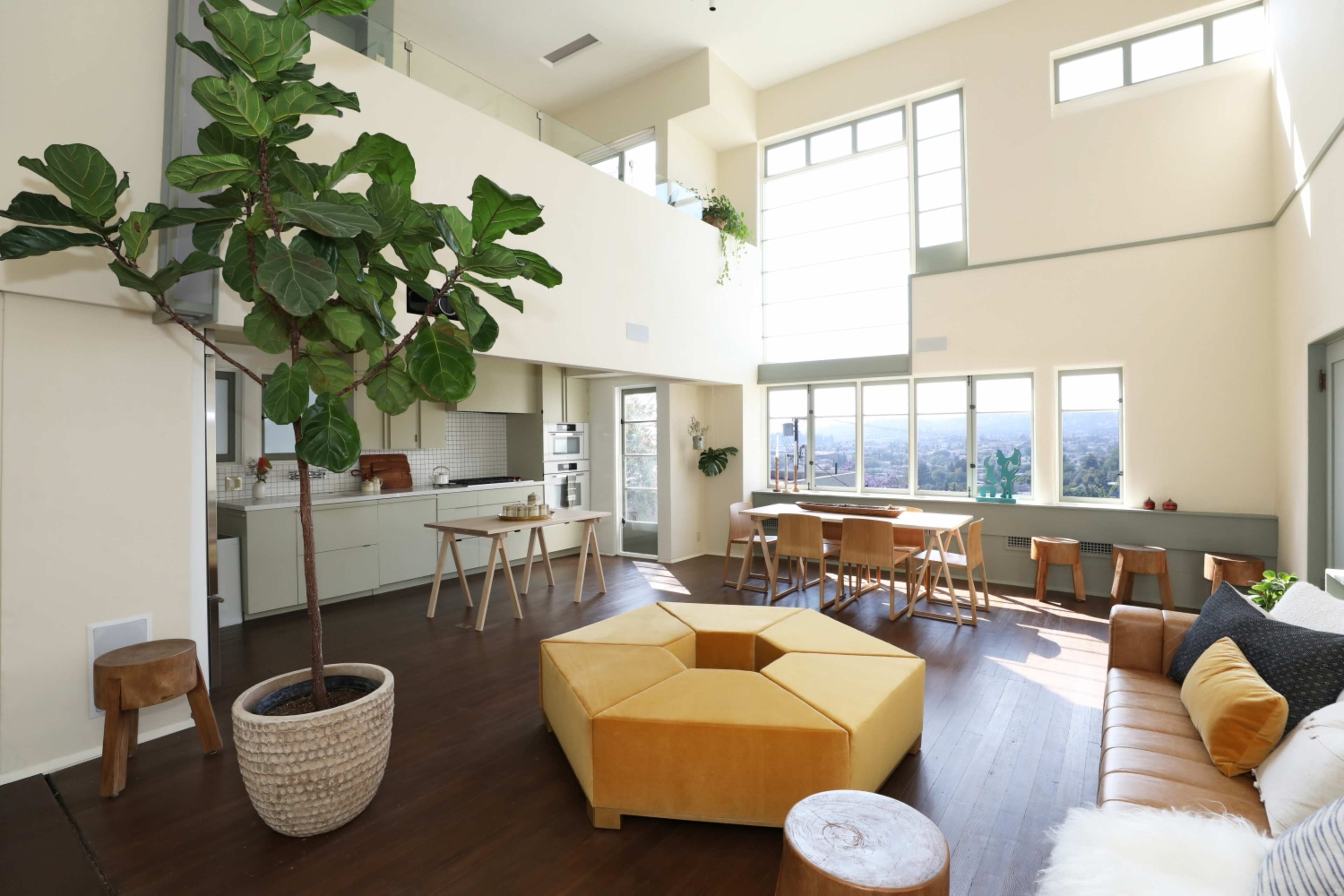 An open-plan living space with large windows, a kitchen area, a dining table, and a seating arrangement featuring a yellow hexagonal ottoman.