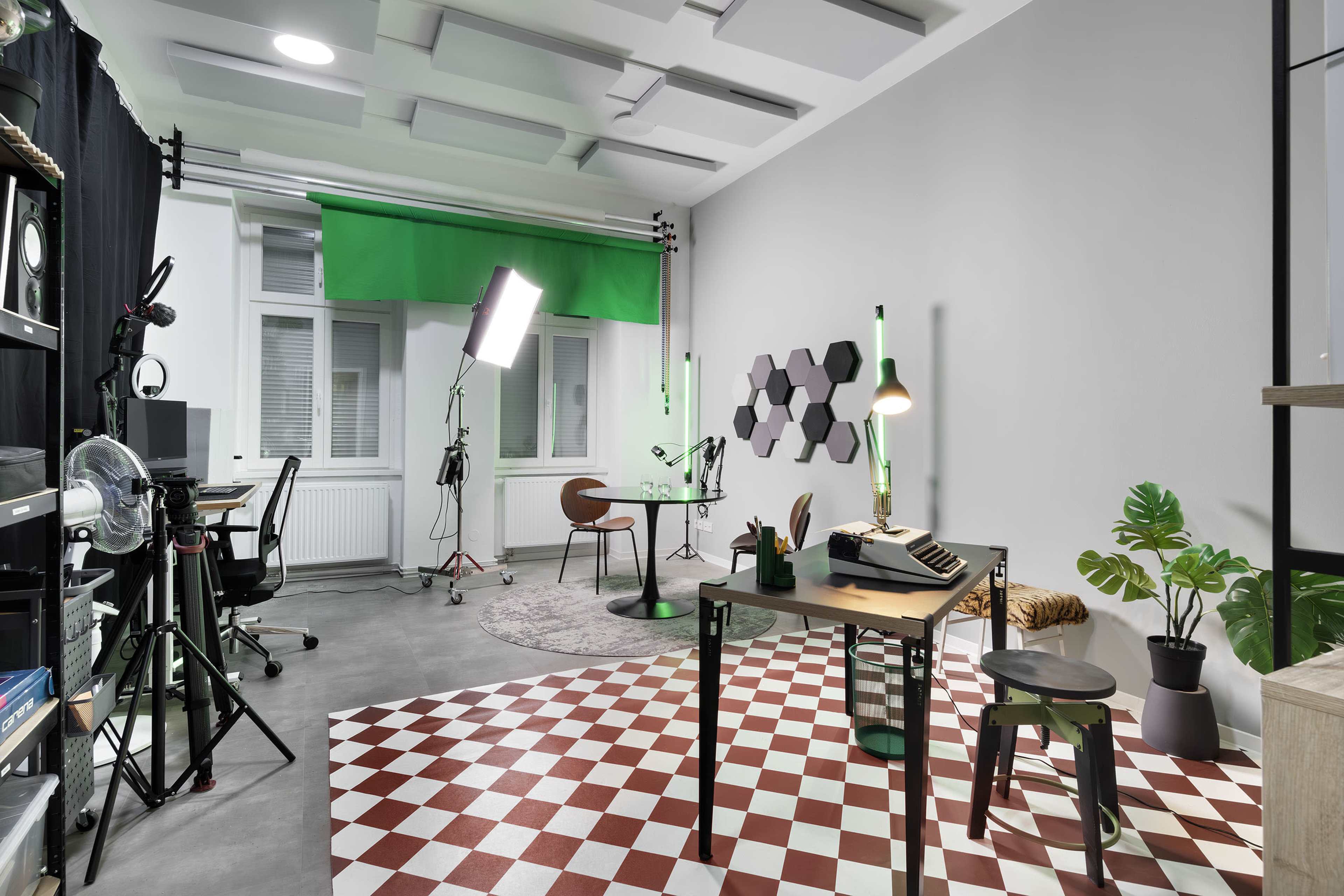 The scene depicts a modern studio workspace with a checkered floor, a desk with a typewriter, a green screen setup, and various potted plants.
