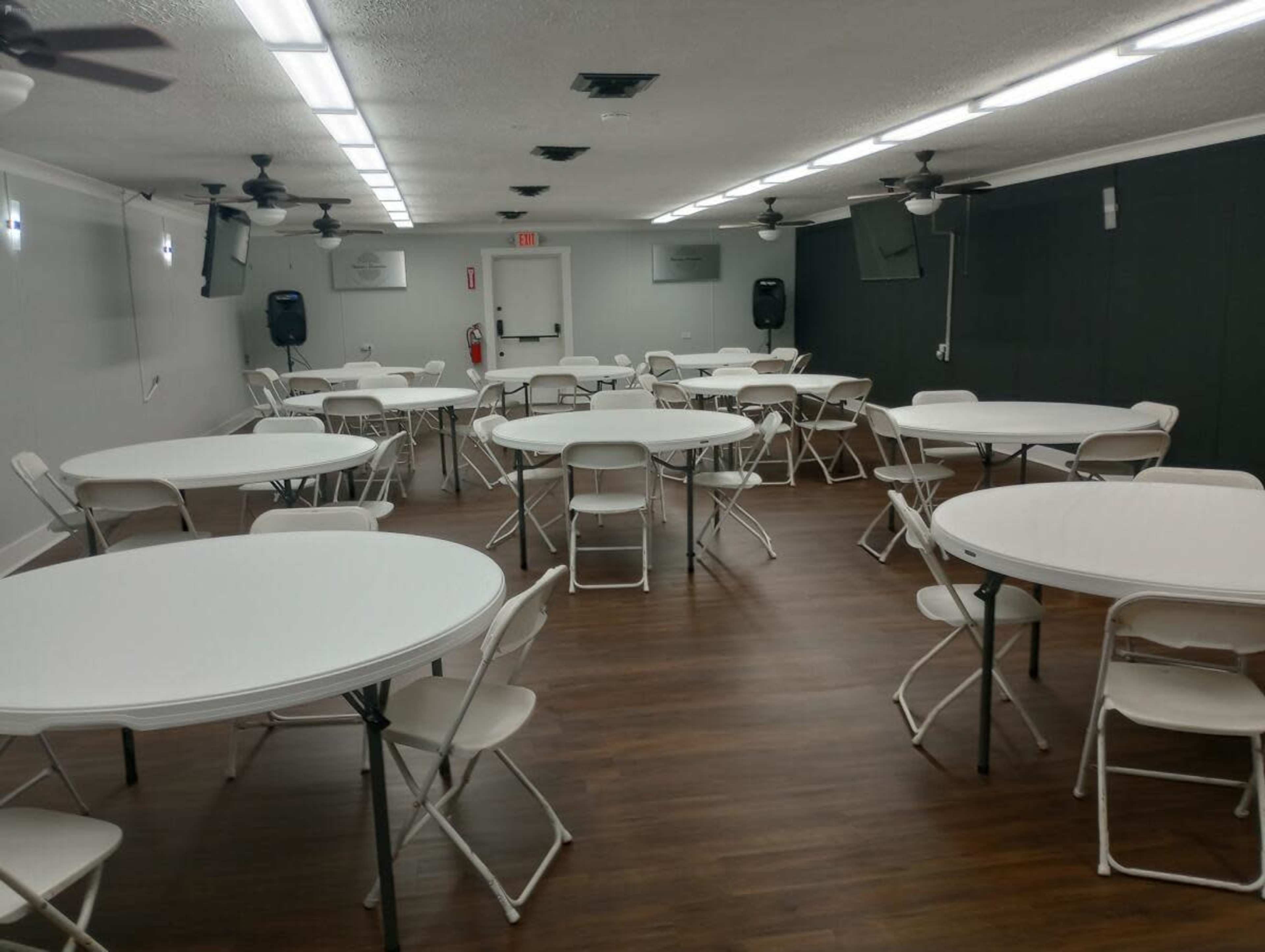 The room features multiple round tables with chairs arranged neatly, under fluorescent lighting and ceiling fans.