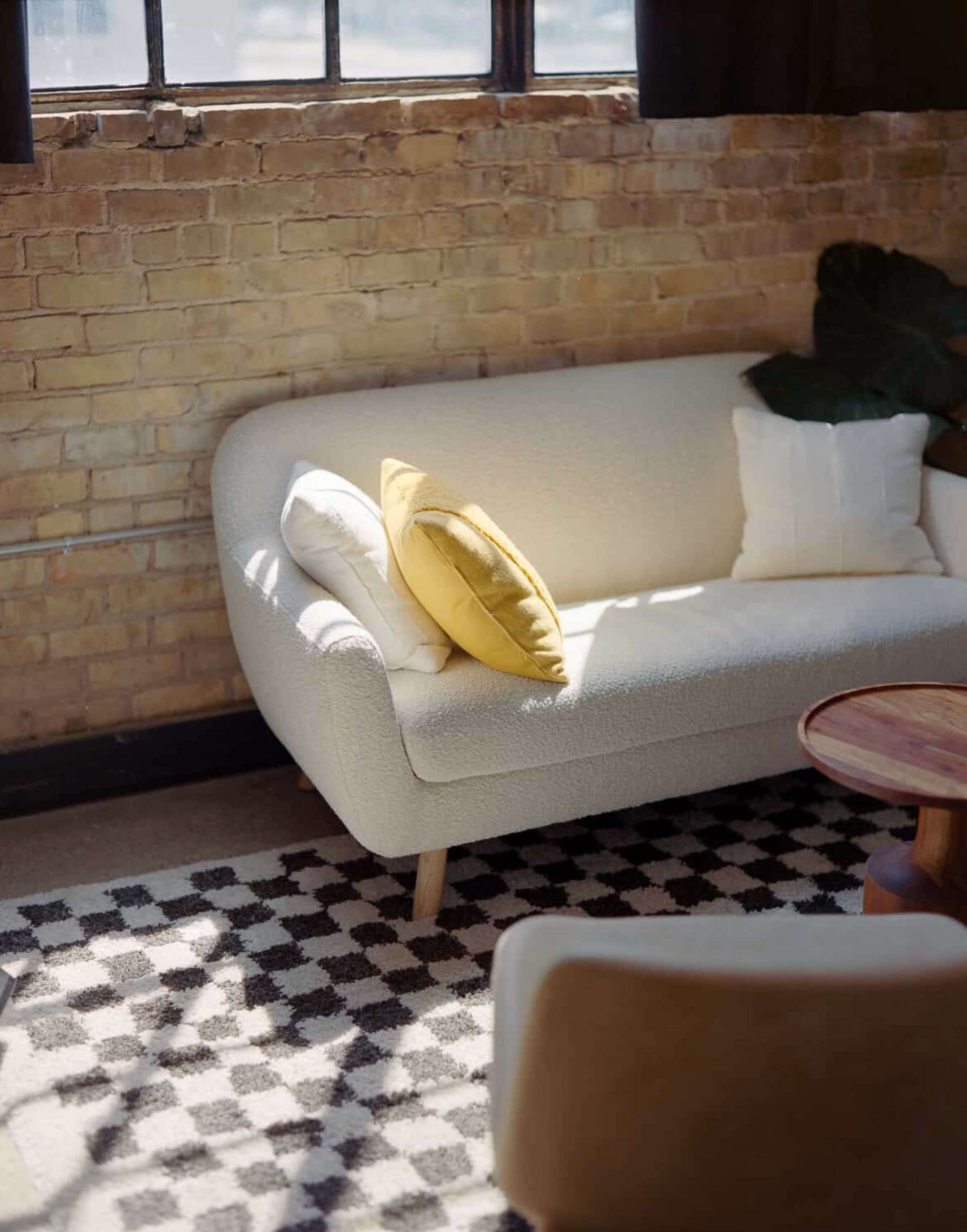A modern light-colored sofa with two pillows sits on a patterned rug beside a wooden coffee table, against a backdrop of exposed brick walls and large windows.