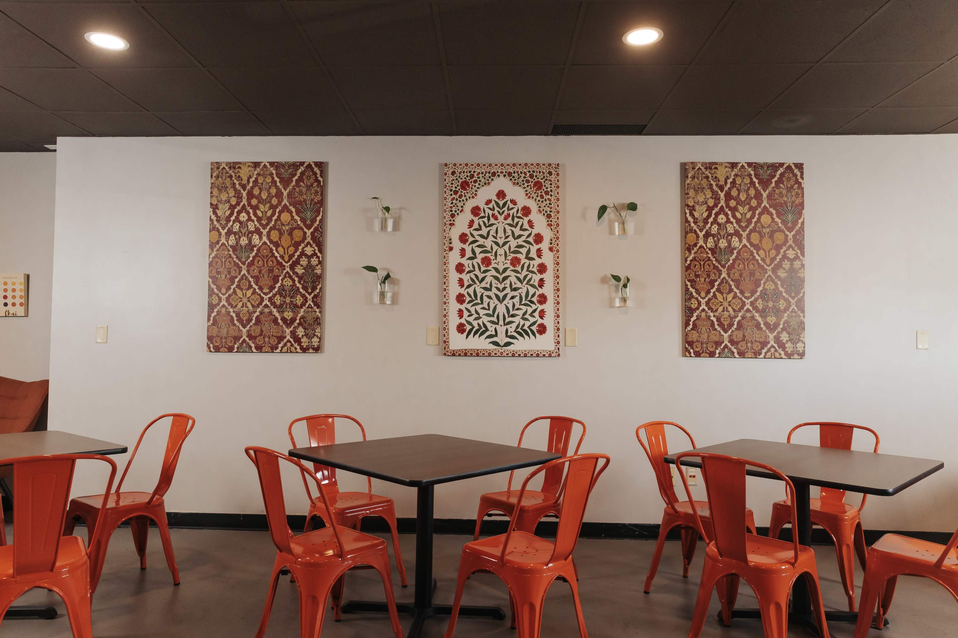 A modern café interior with orange chairs around black tables, featuring decorative wall art and small plants in hanging containers.