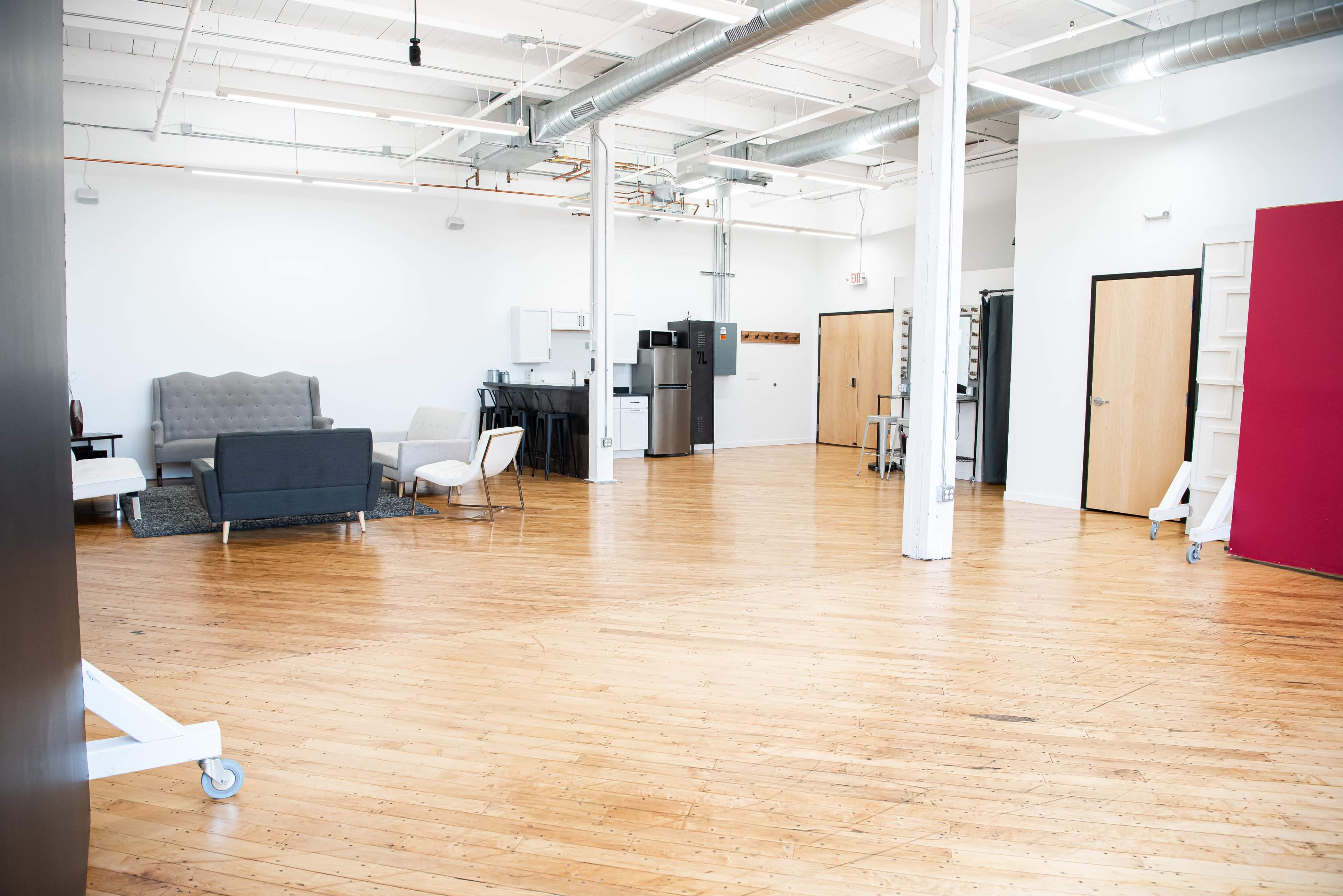 A spacious, empty room with wooden flooring, featuring a few pieces of furniture including a sofa, chairs, and kitchen appliances along one wall.