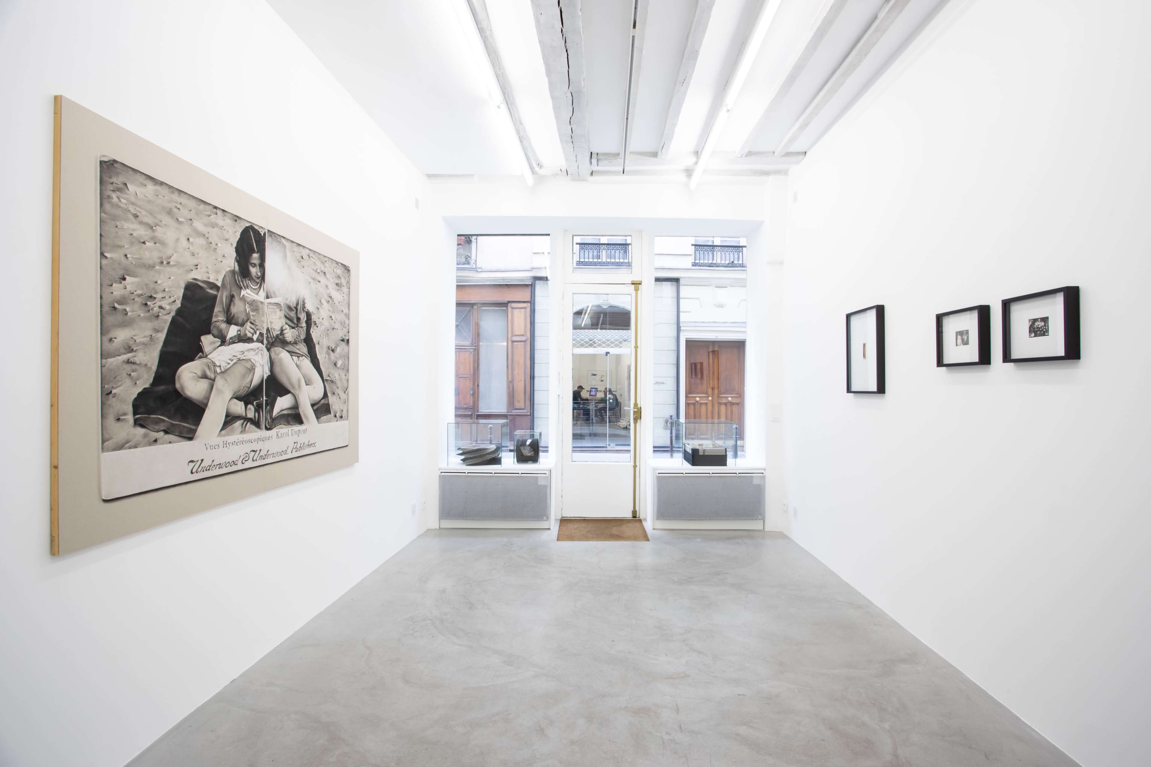 The image shows a minimalistic art gallery featuring a large black-and-white photograph on the left wall and several smaller framed artworks on the right.