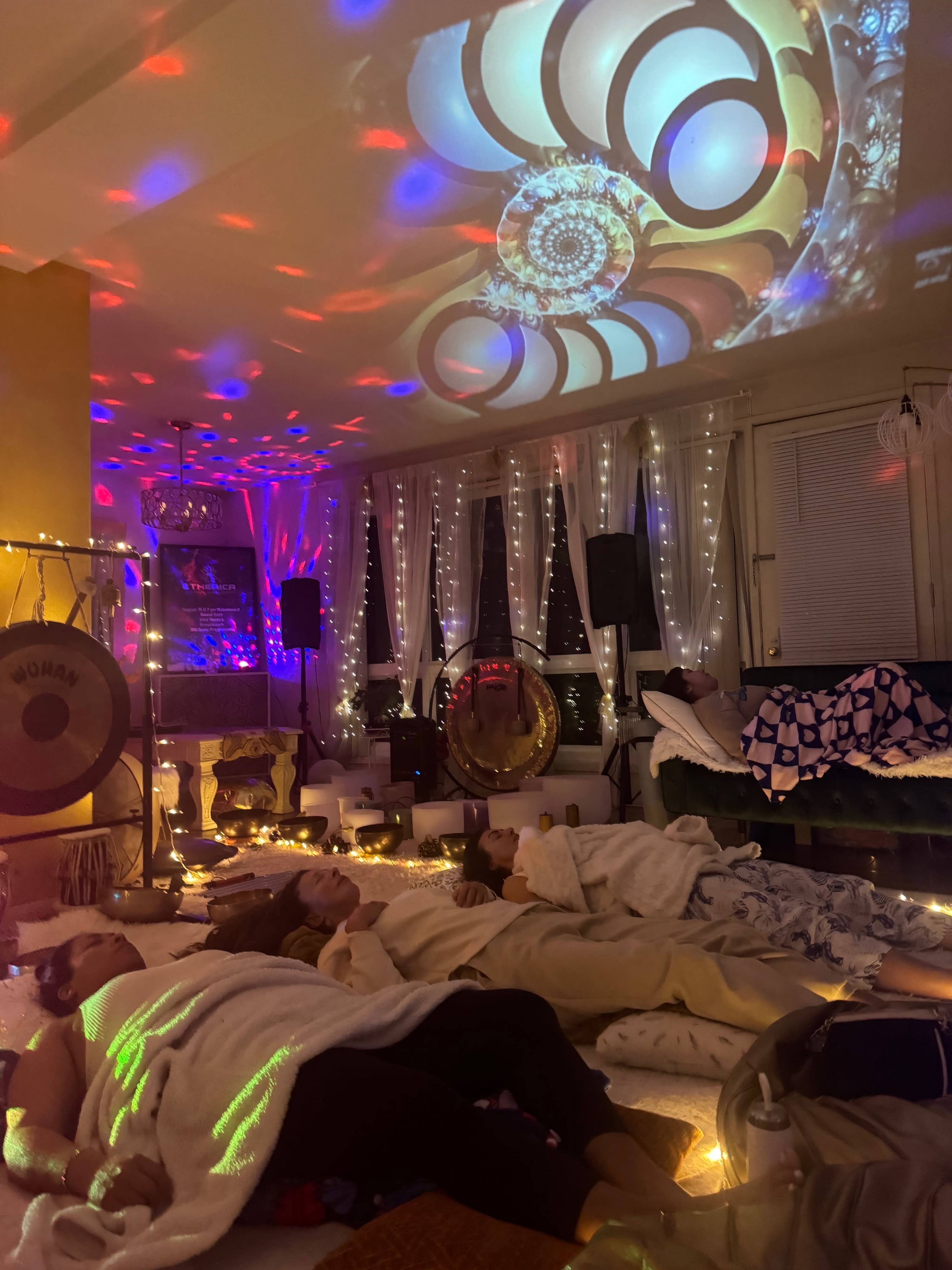 A group of people lies on the floor of a room adorned with colorful lights, projected patterns, and cozy blankets, while various instruments are arranged in the background.