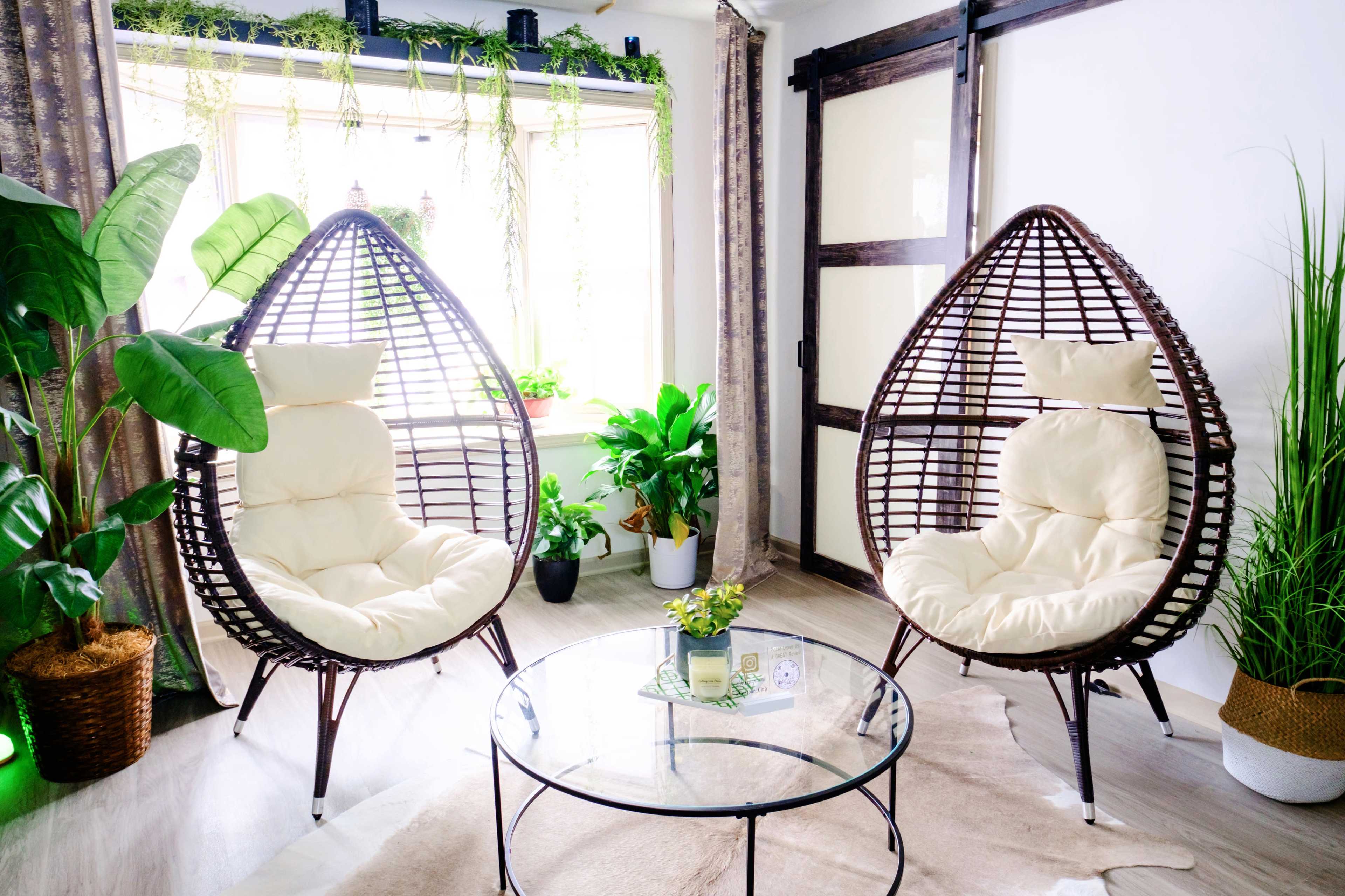 A cozy seating area featuring two egg-shaped chairs with cream cushions, positioned beside a glass coffee table and surrounded by various indoor plants.