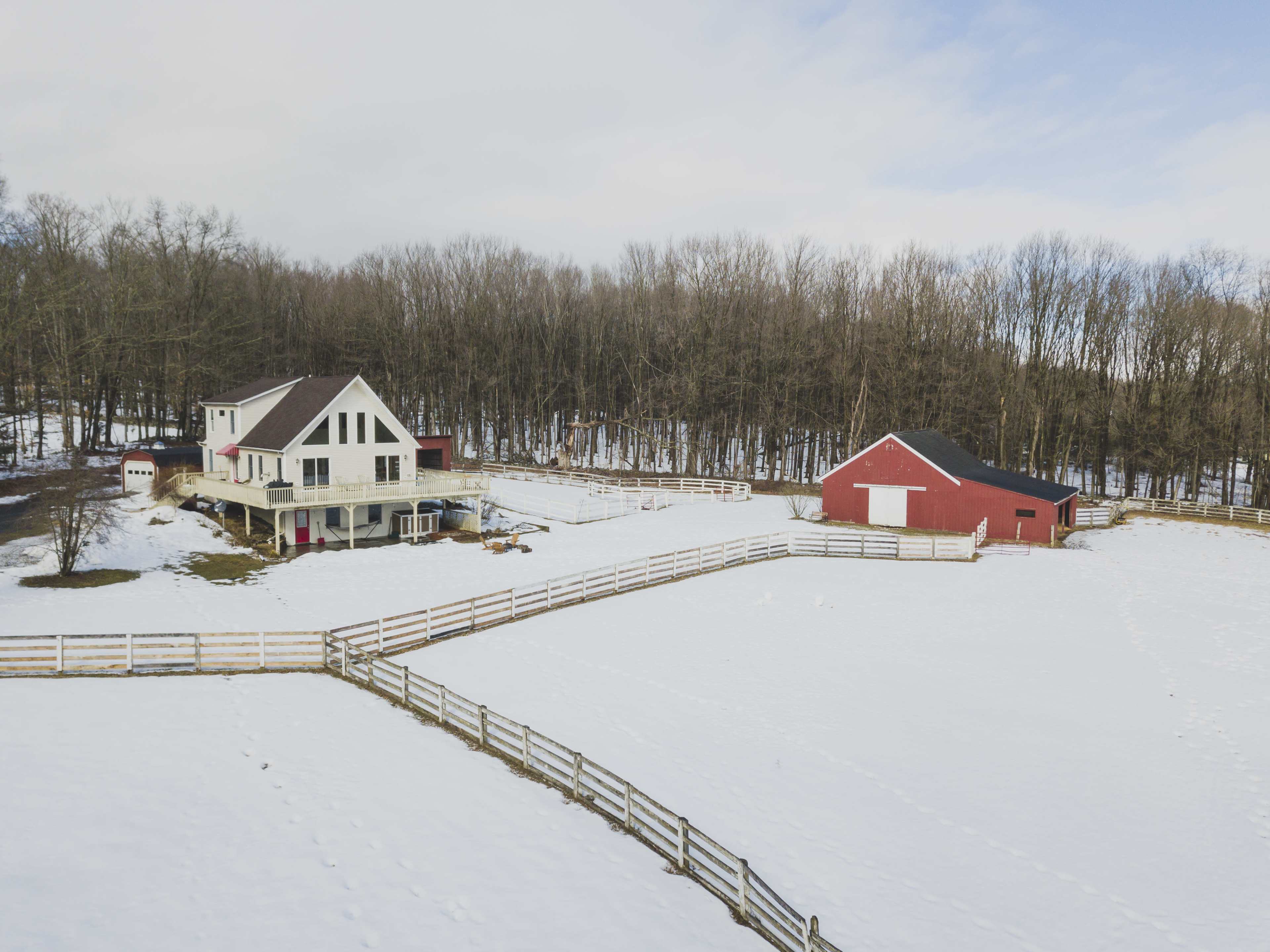 A snowy landscape features a large farmhouse with a sloped roof and a red barn, surrounded by a wooden fence and wooded area in the background.
