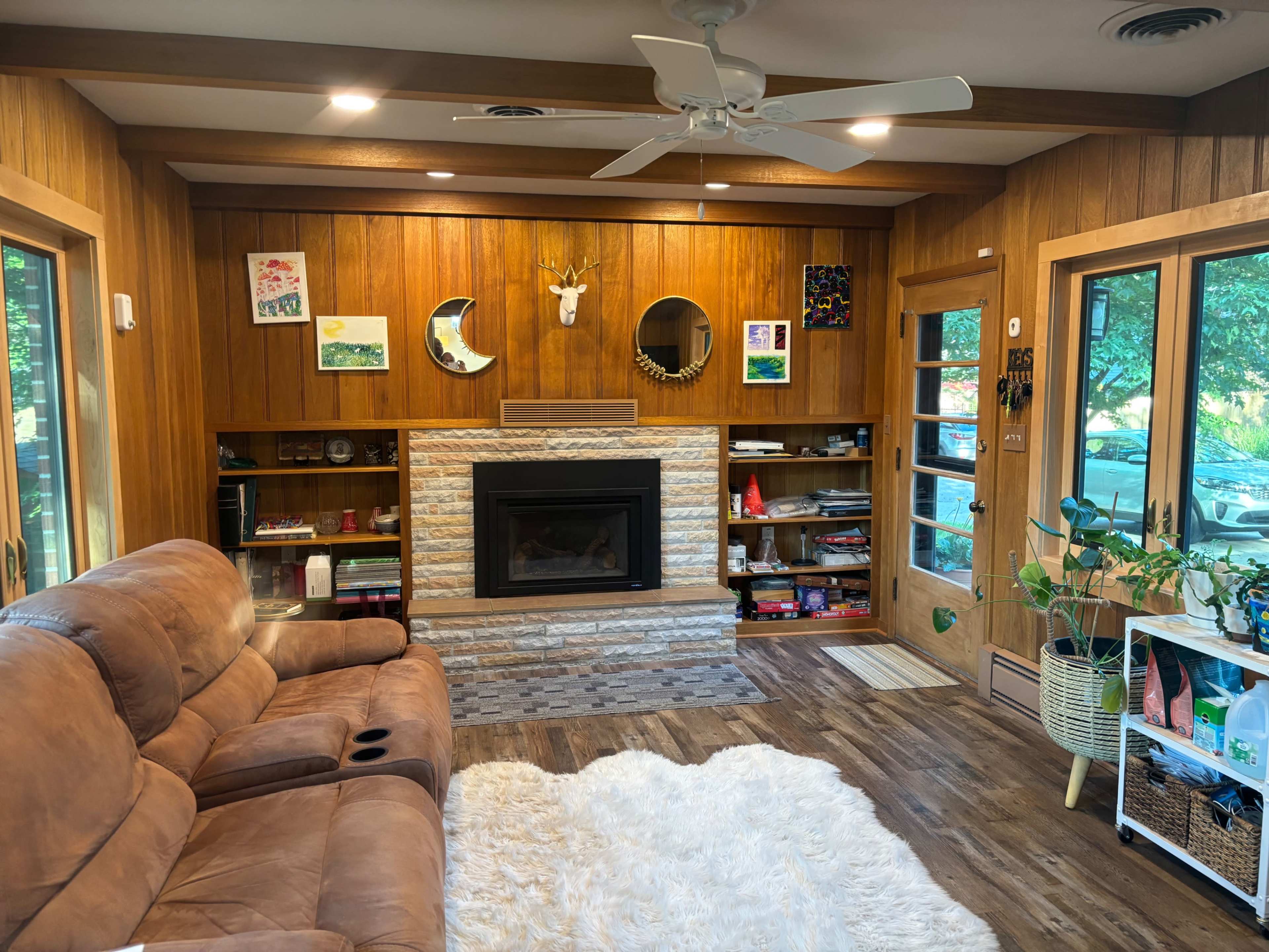 The image shows a cozy living room with a brown sofa, a stone fireplace, wooden paneling, and decorative artwork on the walls.