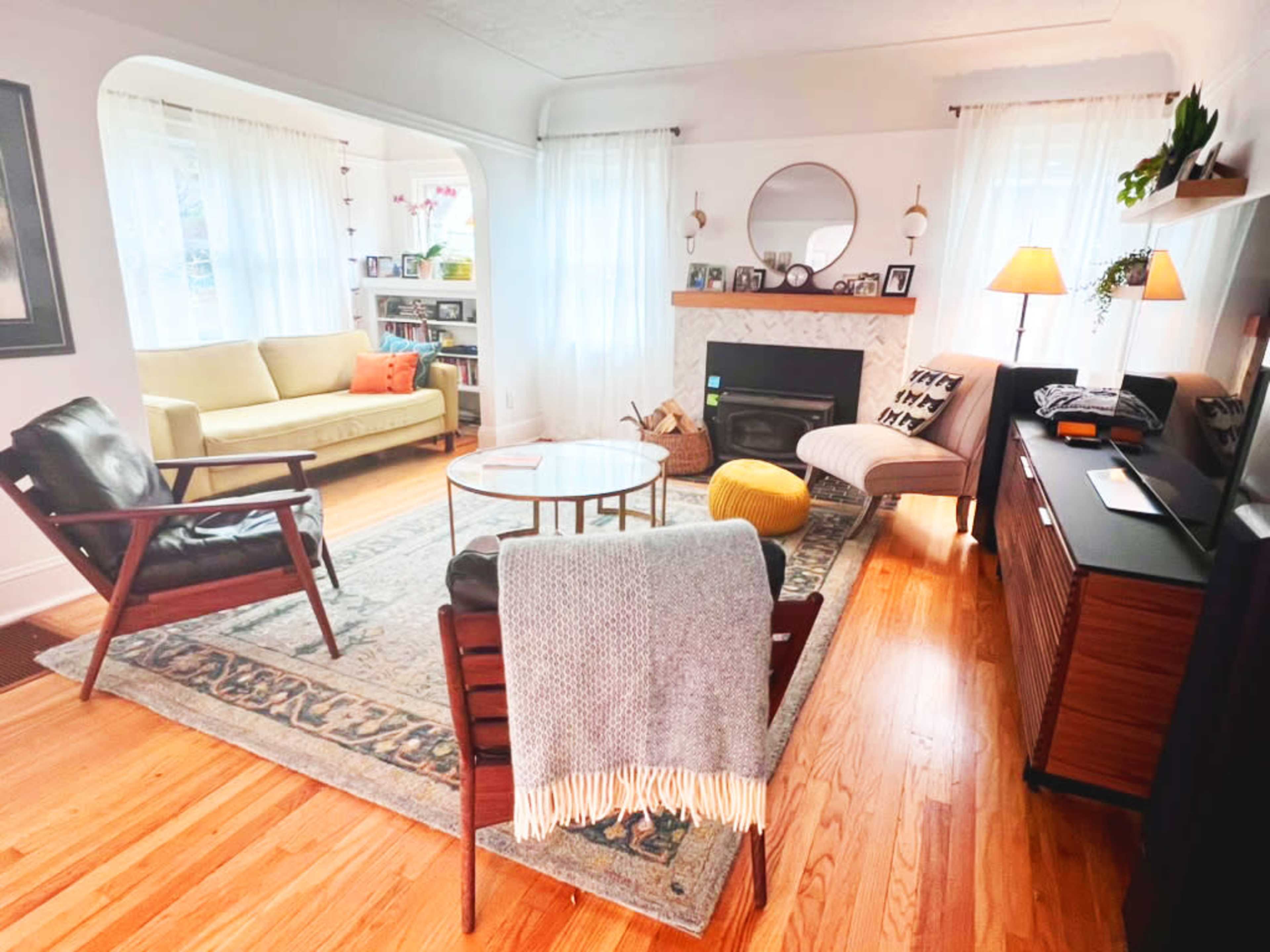 The living room features a sofa, two armchairs, a coffee table, and a fireplace with a mantel, surrounded by large windows and a patterned rug.