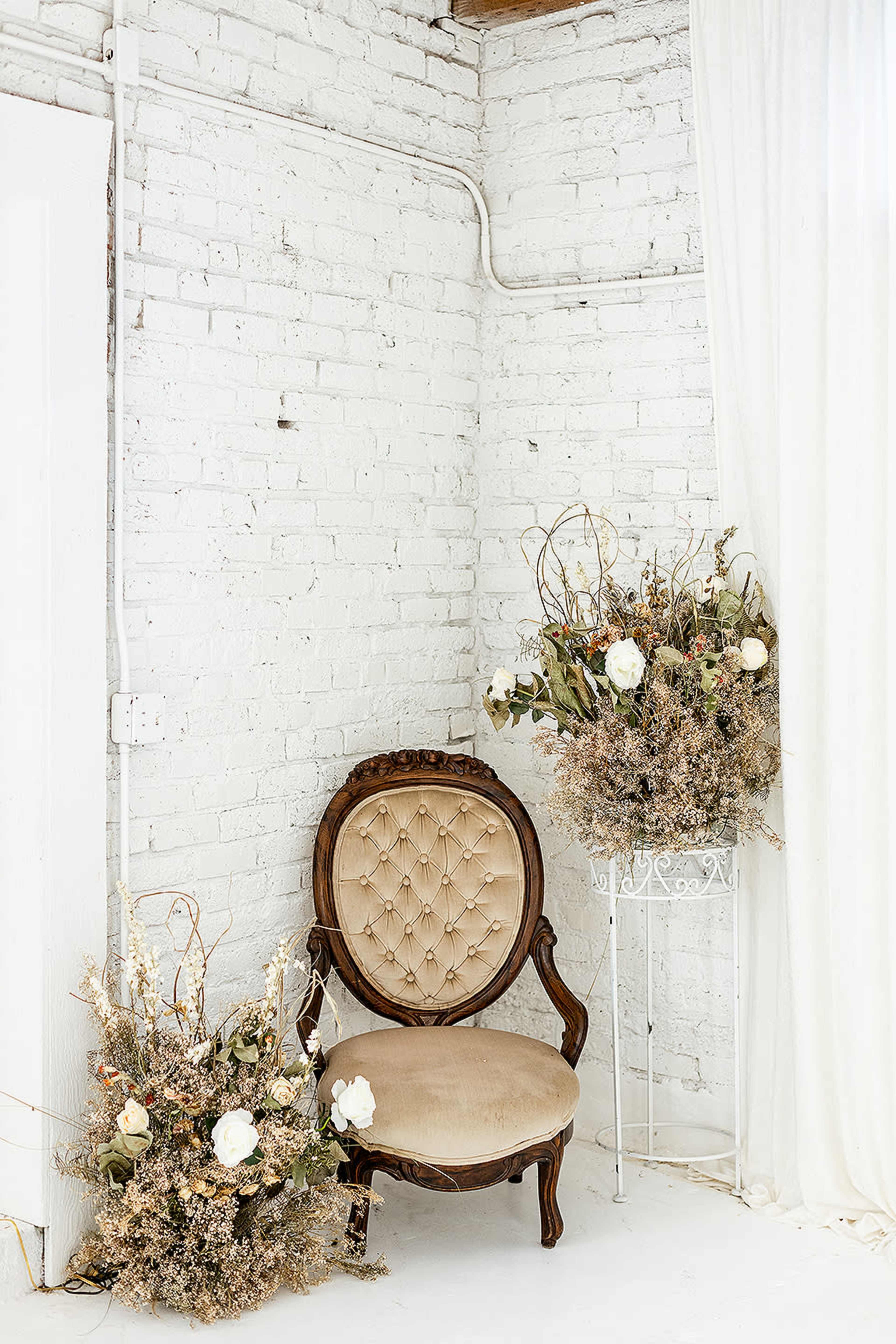 A vintage upholstered chair is placed in a corner next to large floral arrangements in decorative containers against a white brick wall.