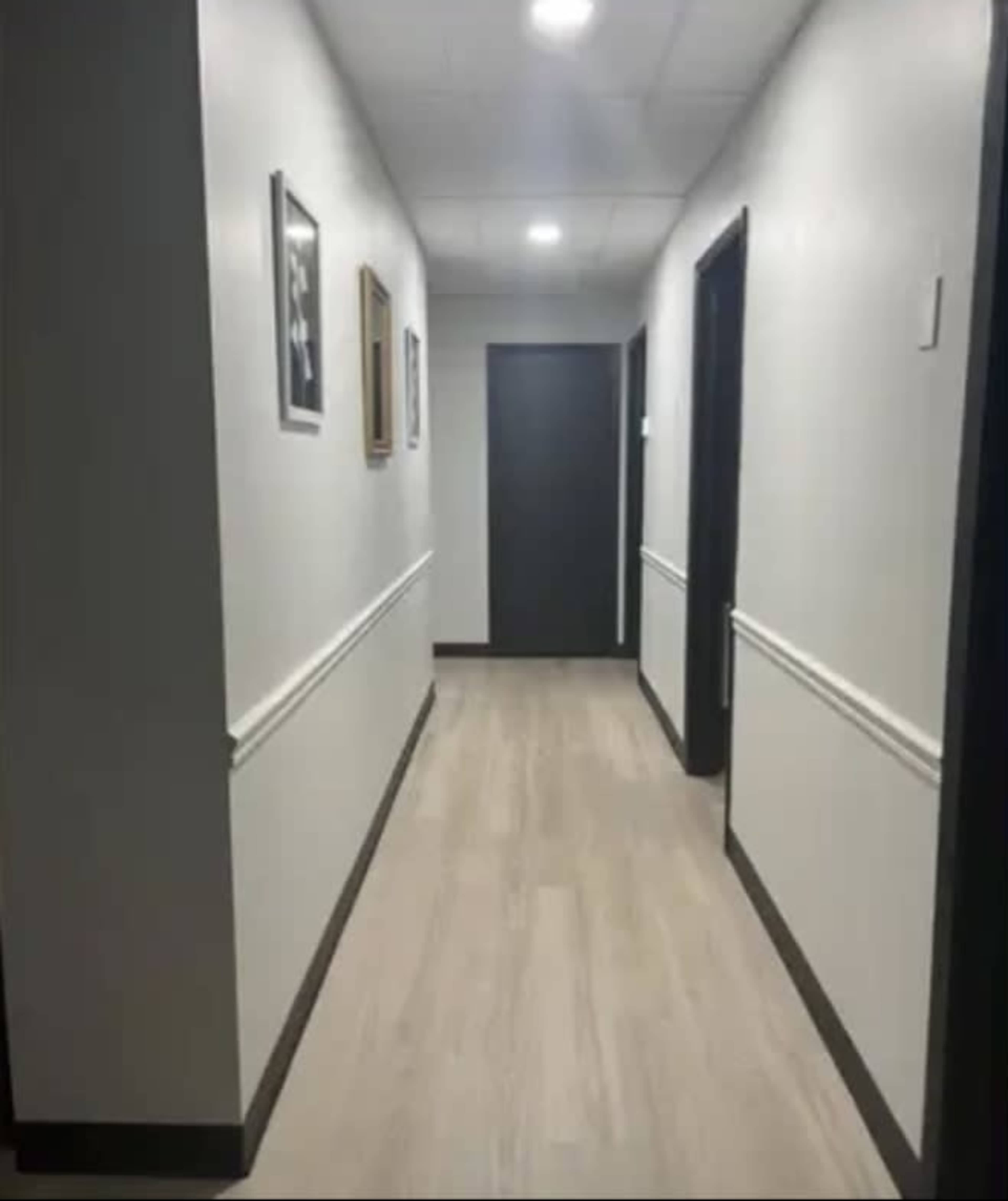 The image shows a narrow hallway with light-colored walls, framed pictures on the left, and a dark door at the end.
