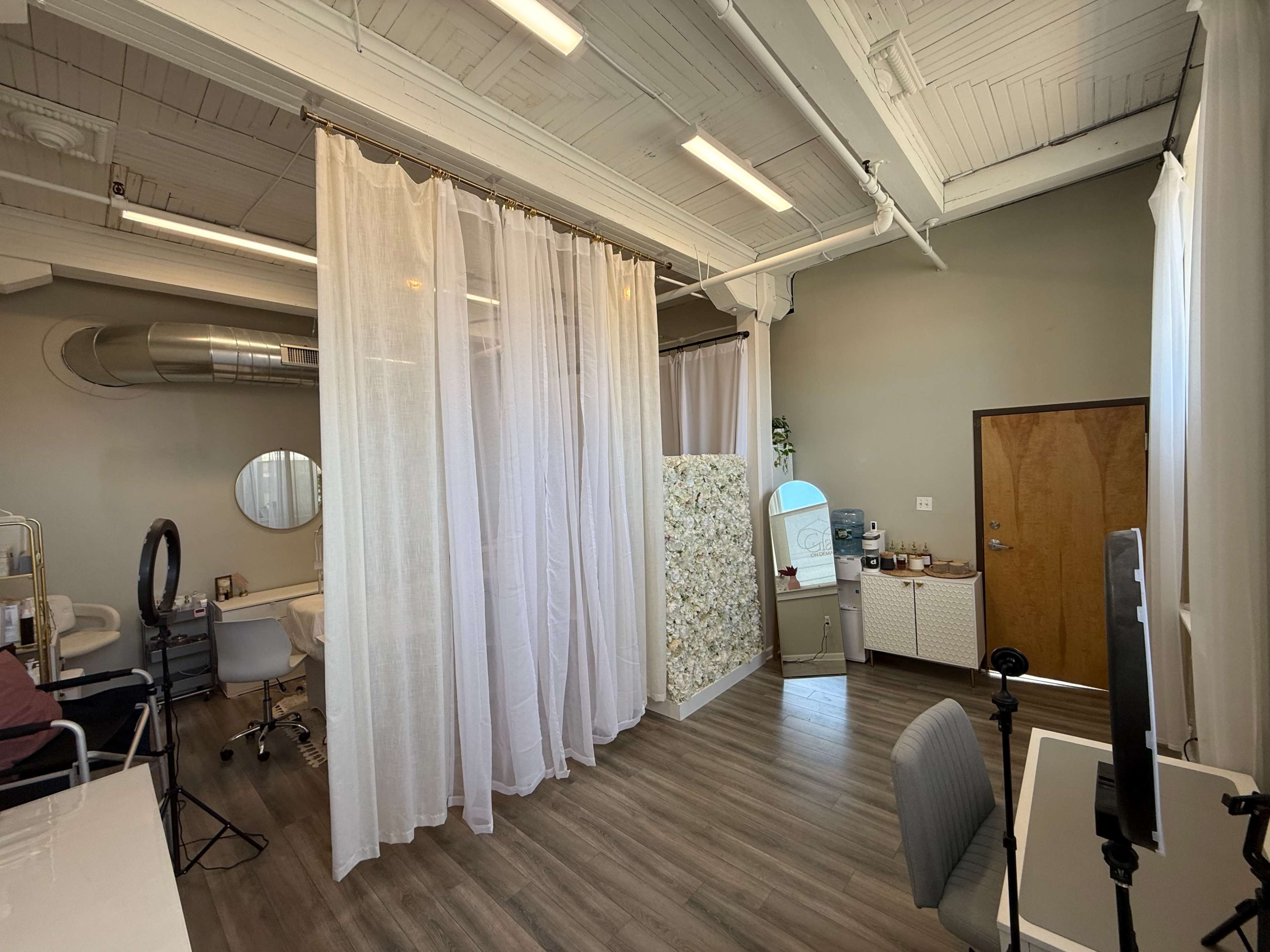 The image shows a bright, open room divided by sheer curtains, featuring a vanity area with a mirror and a door leading outside.