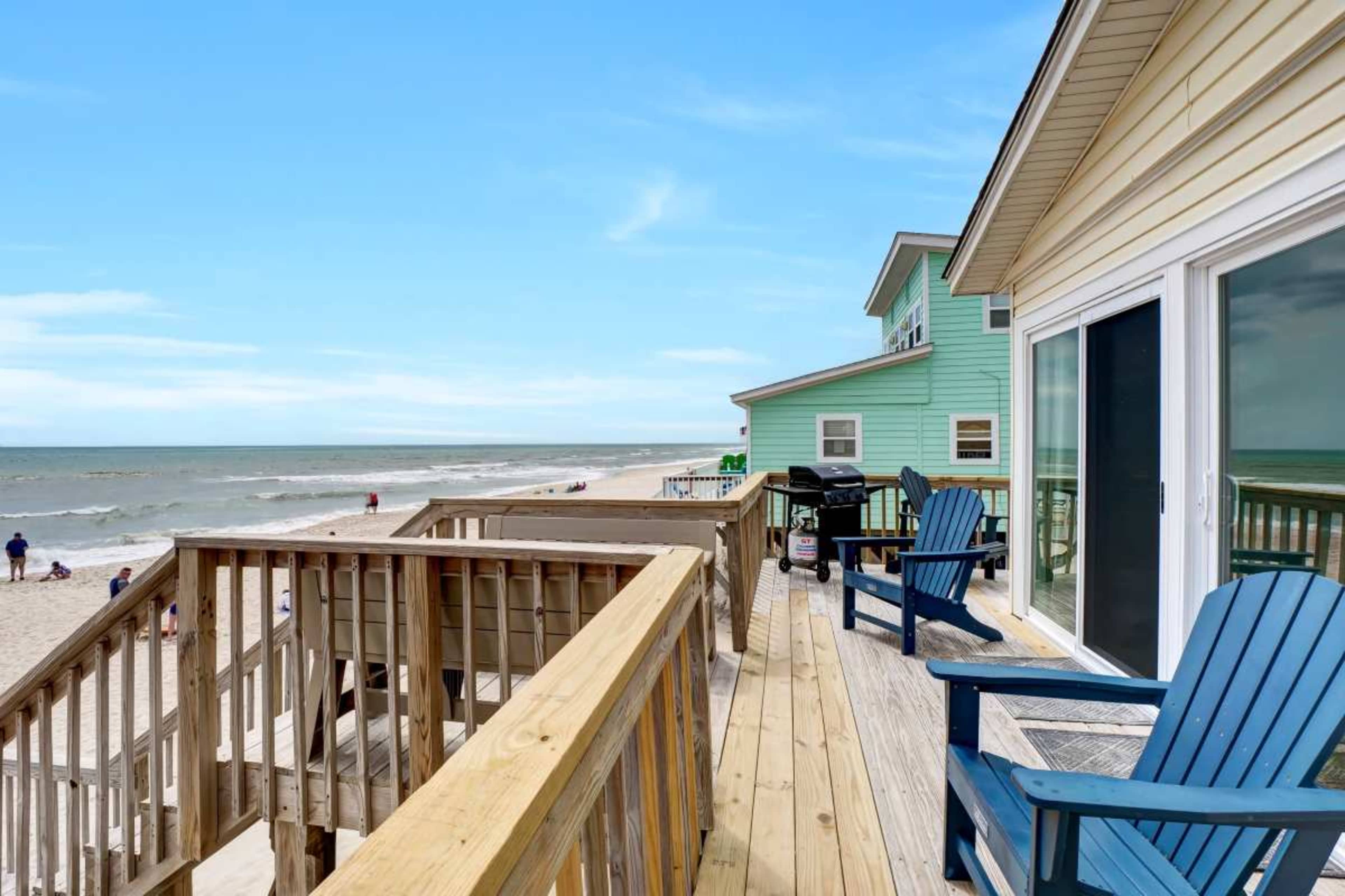 A wooden deck with blue chairs overlooks a sandy beach and the ocean, while nearby houses are painted in vibrant colors.