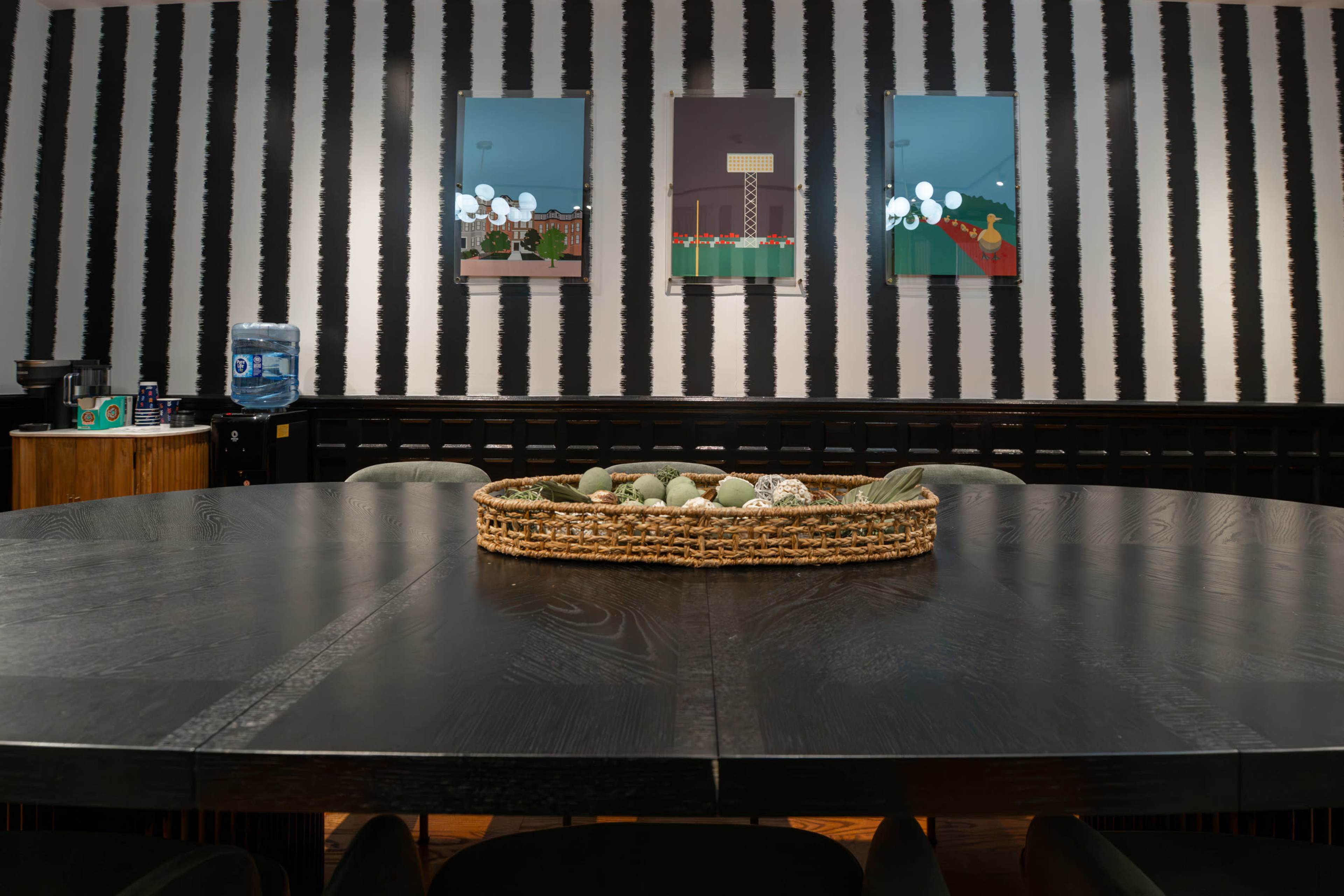 A large round wooden table with a woven basket containing decorative items sits in a room featuring striped walls and two framed artworks.