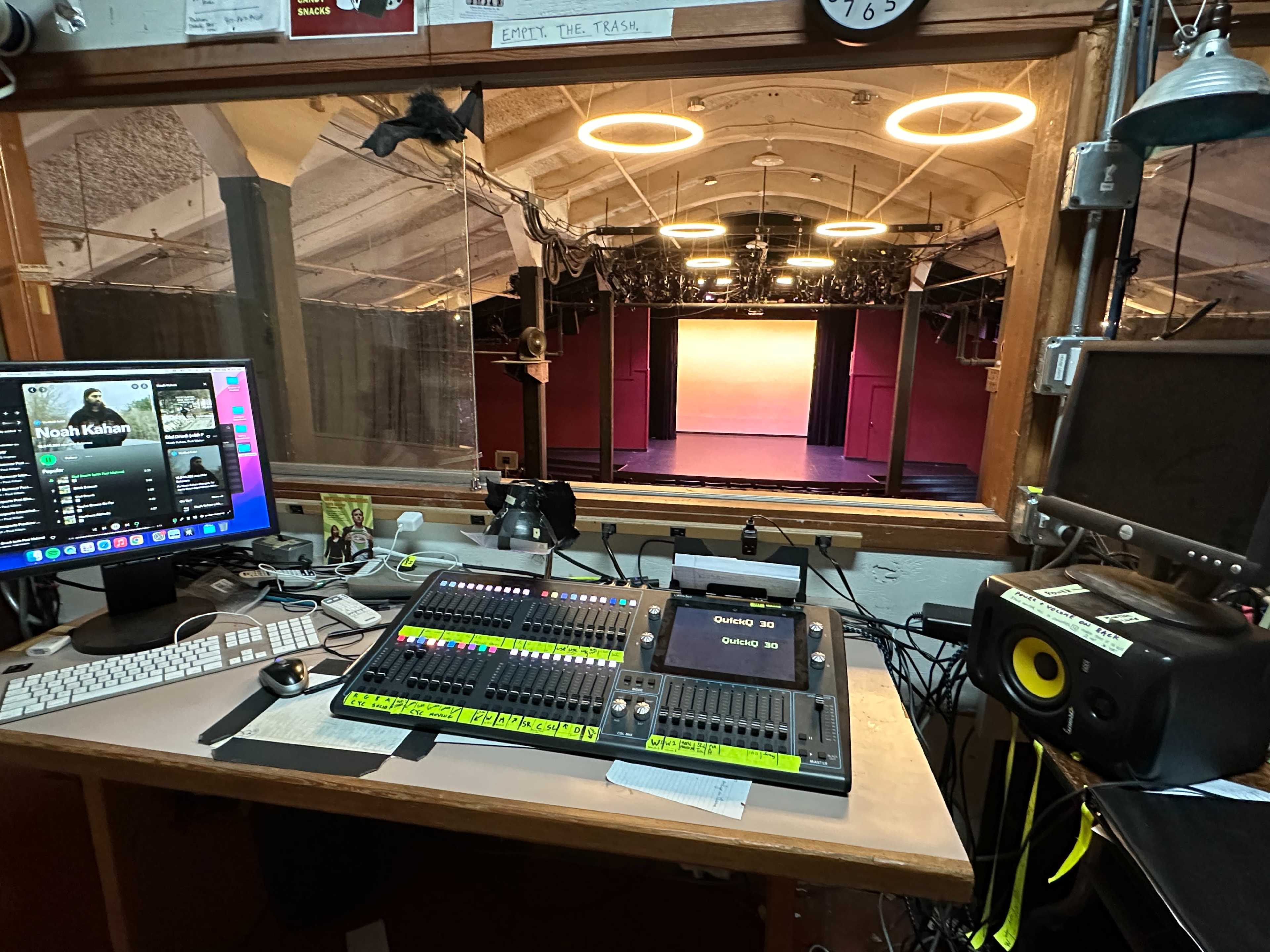 The image shows a control room with a soundboard and computer, overlooking a dimly lit stage with colorful lighting.
