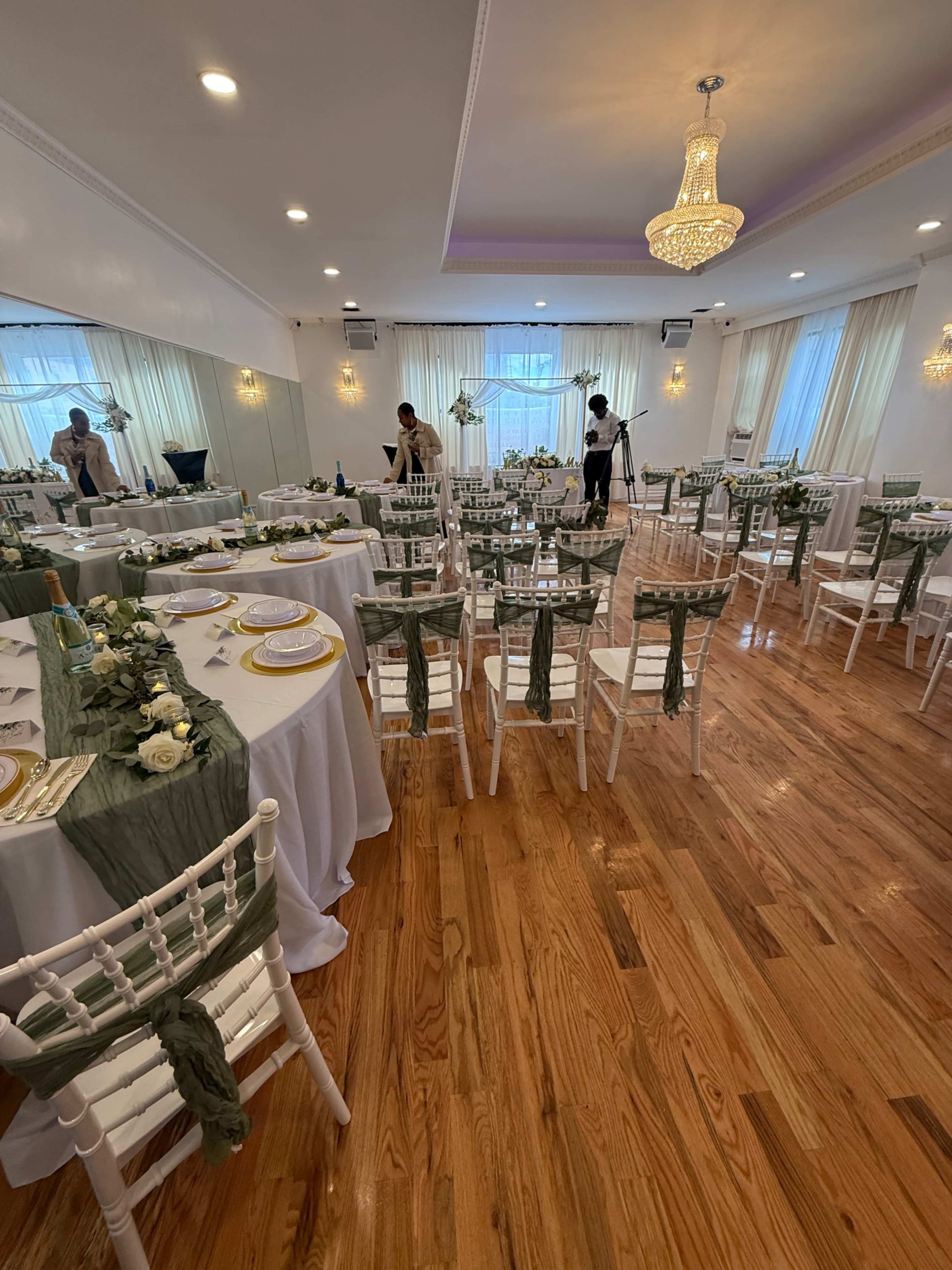 The image shows a banquet hall set up for an event with multiple round tables, white chairs draped in green fabric, and floral decorations.