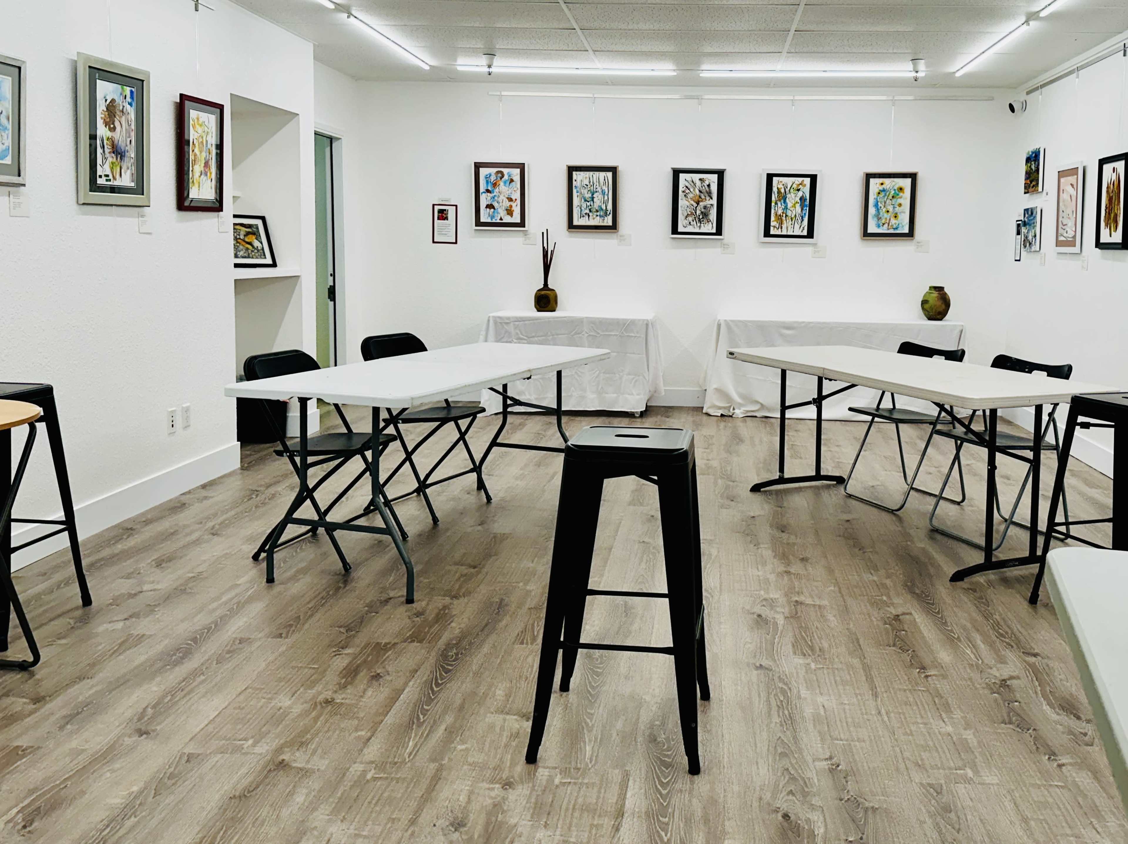 A spacious art gallery with several folding tables and black chairs arranged around the room, featuring framed artworks on the white walls.